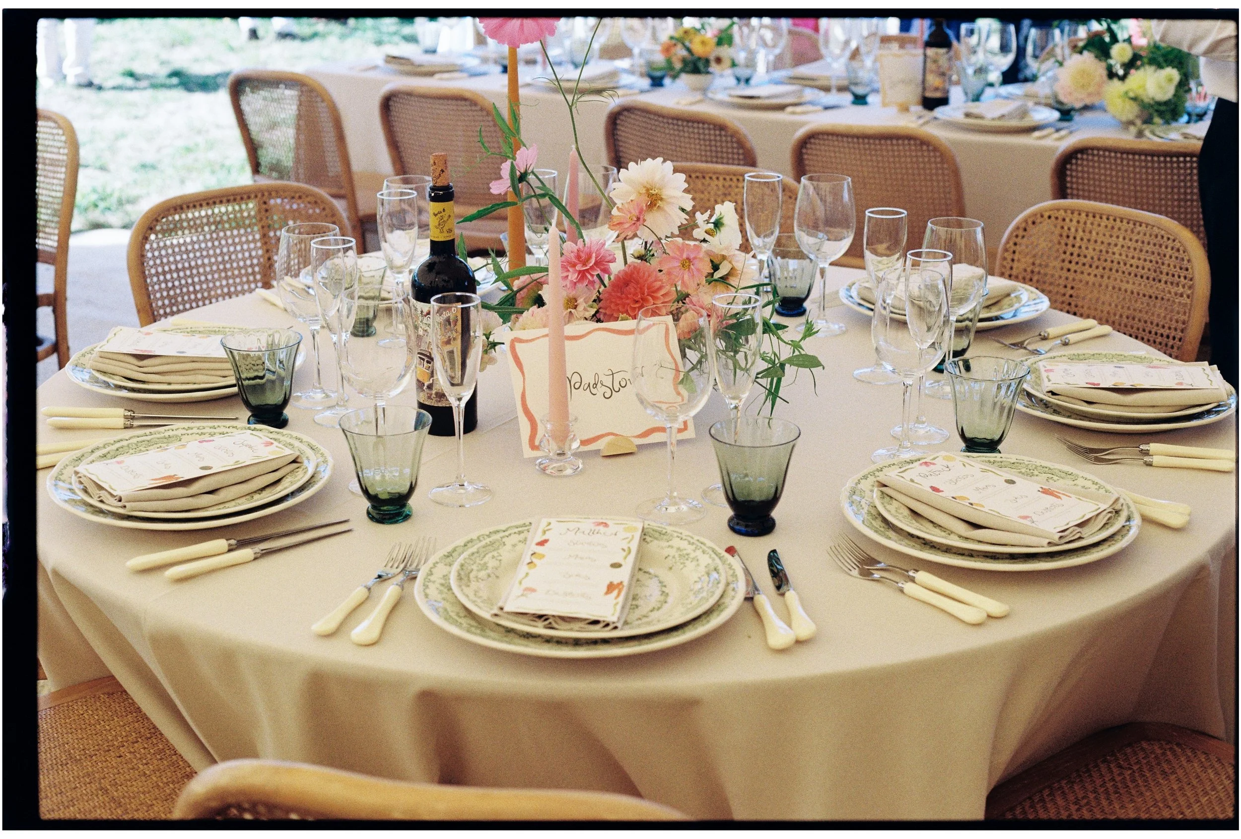 Round banquet table set for a celebration, decorated with pink and white flowers, candles, wine, and place settings with napkins and menus in an outdoor or semi-outdoor setting.