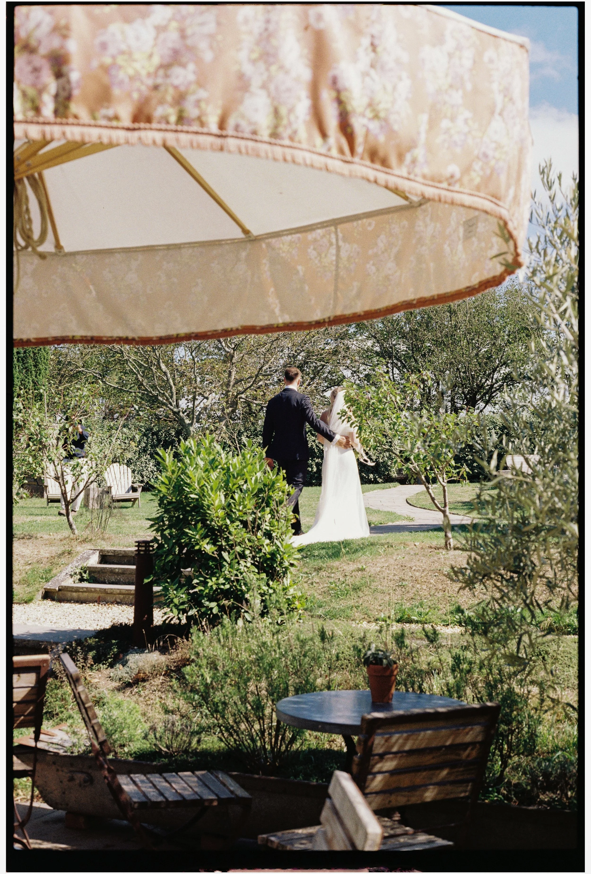 A couple dressed in wedding attire walking hand in hand through a lush garden, taken from underneath a floral-patterned patio umbrella, with outdoor furniture and trees in the background.