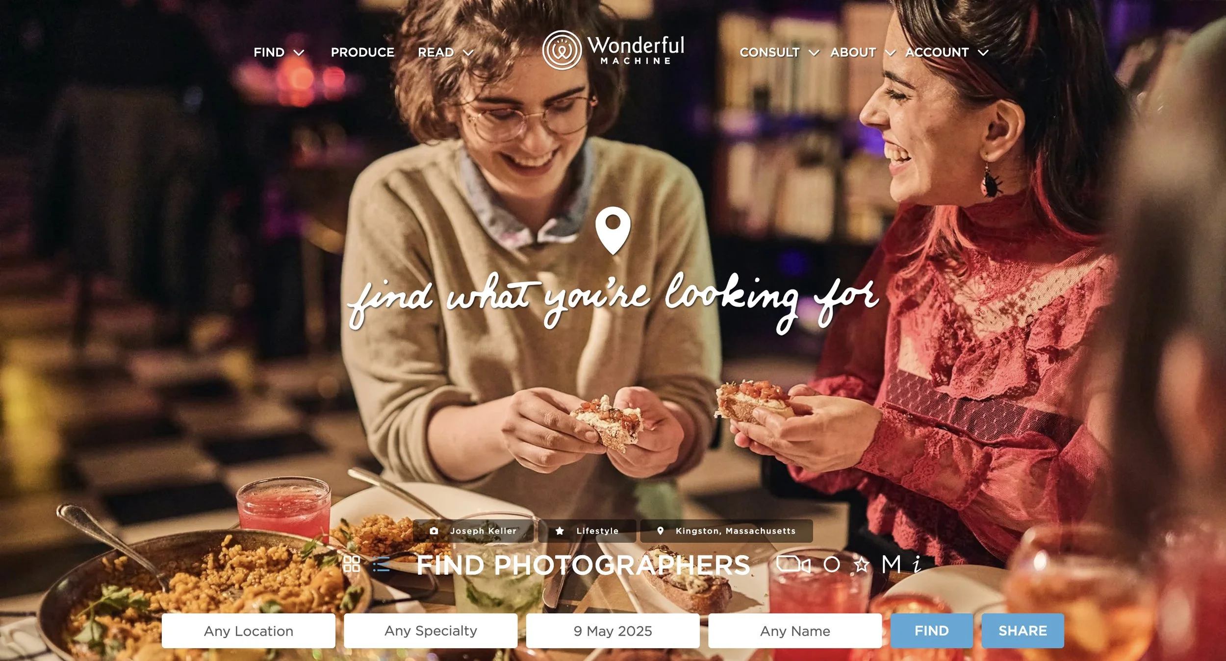 Two women are smiling and enjoying food at a table in a cozy, dimly lit setting. The table has various dishes and drinks, and the background features bookshelves and warm lighting.
