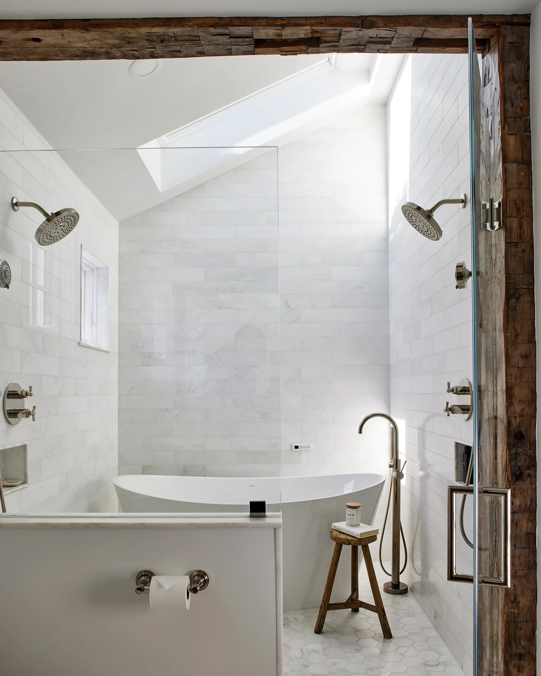 Modern white bathroom with a bathtub, two showerheads, a small window, and a skylight ceiling. Wooden accents around the shower area and a small wooden stool with a candle and book.