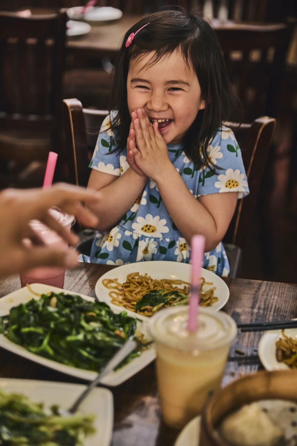 A young girl with dark hair, wearing a blue dress with a daisy pattern, is happily laughing and clapping her hands together while sitting at a restaurant table with various dishes, including noodles, leafy greens, and drinks with pink straws.