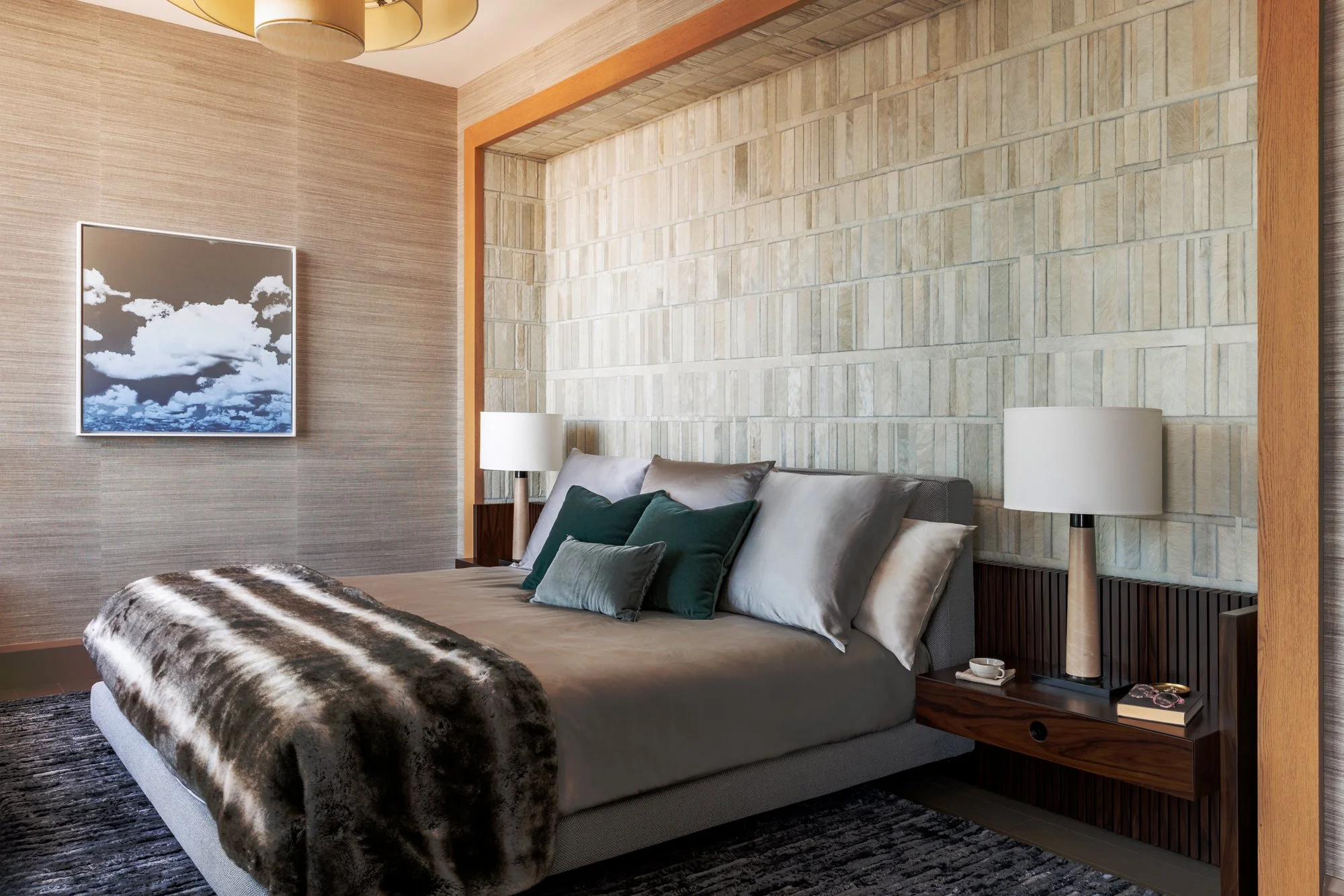 A bedroom with a neatly made bed with multiple pillows, a fur throw at the foot, and a dark wooden side table with a lamp, cup, and book. The room features a textured wall behind the bed, a framed picture of clouds on the adjacent wall, and soft ligh