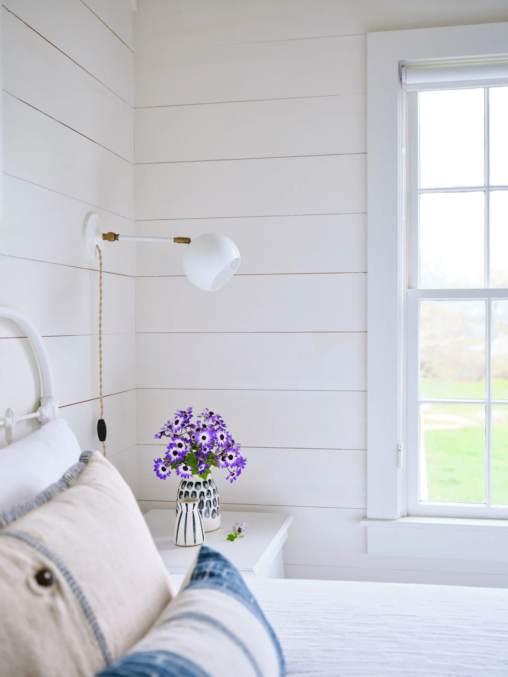 Bedroom with white wooden walls, a window, a white nightstand with a vase of purple flowers, and a bed with pillows and a blanket. Boston based interiors and architectural photographer.  Cottage style.