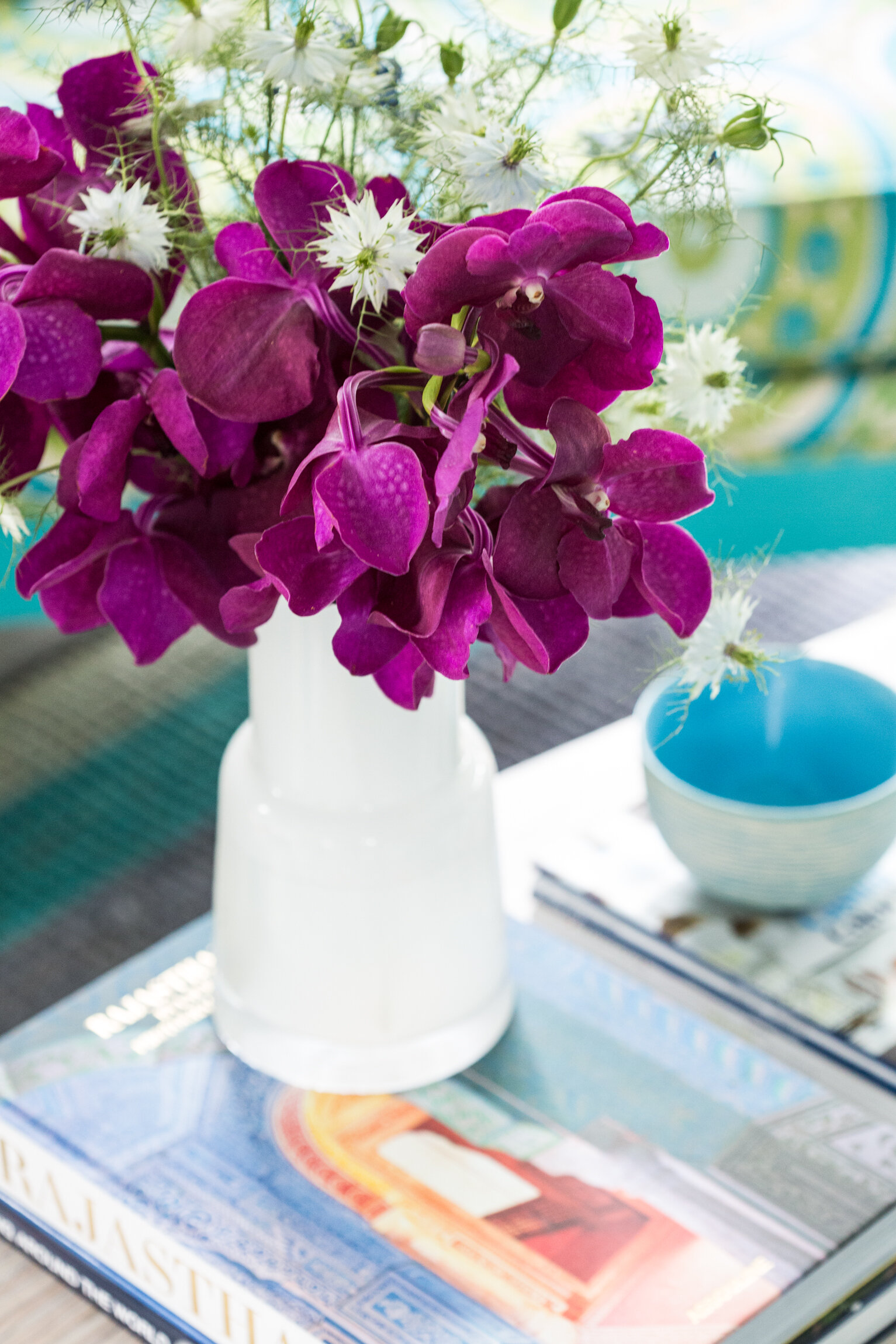 Vase with purple orchids, white flowers, and greenery on top of a magazine on a table, with a blue bowl nearby.