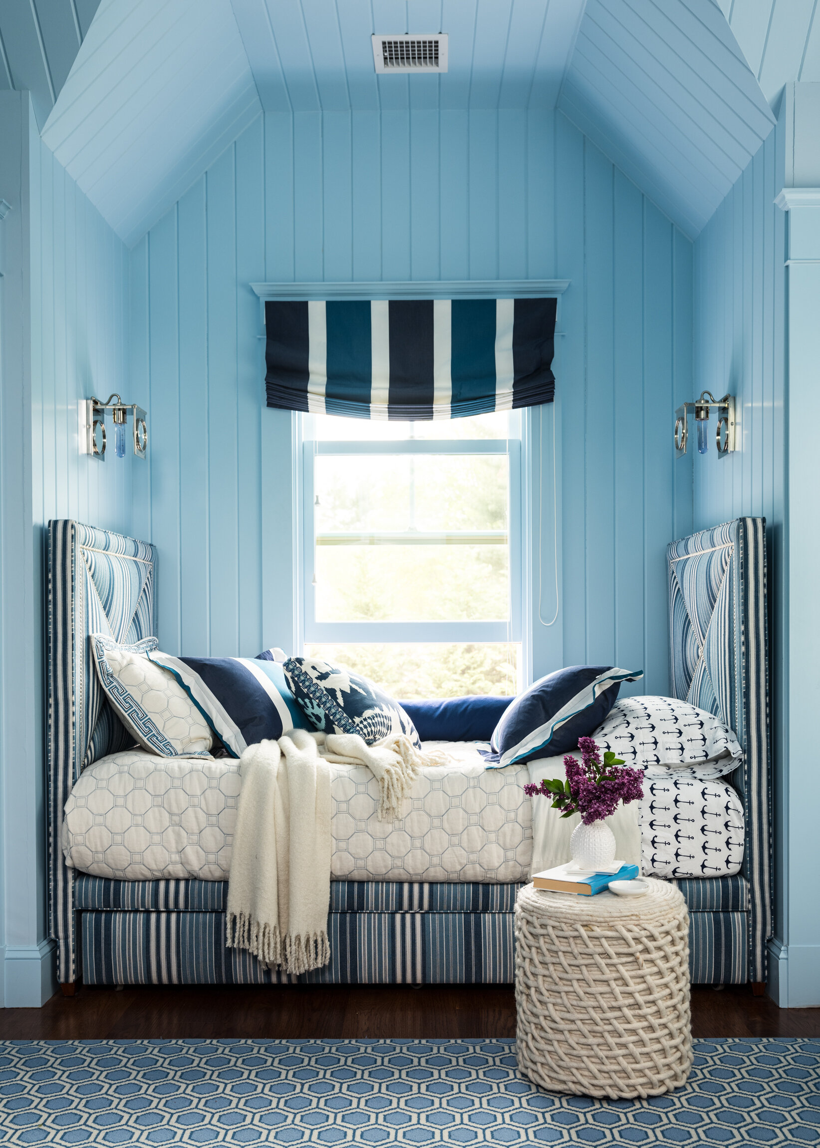 A cozy bedroom with a blue color scheme, featuring a daybed with nautical-themed pillows, a white blanket, a vases of purple flowers, and a patterned blue rug. Boston based interiors and architectural photographer.