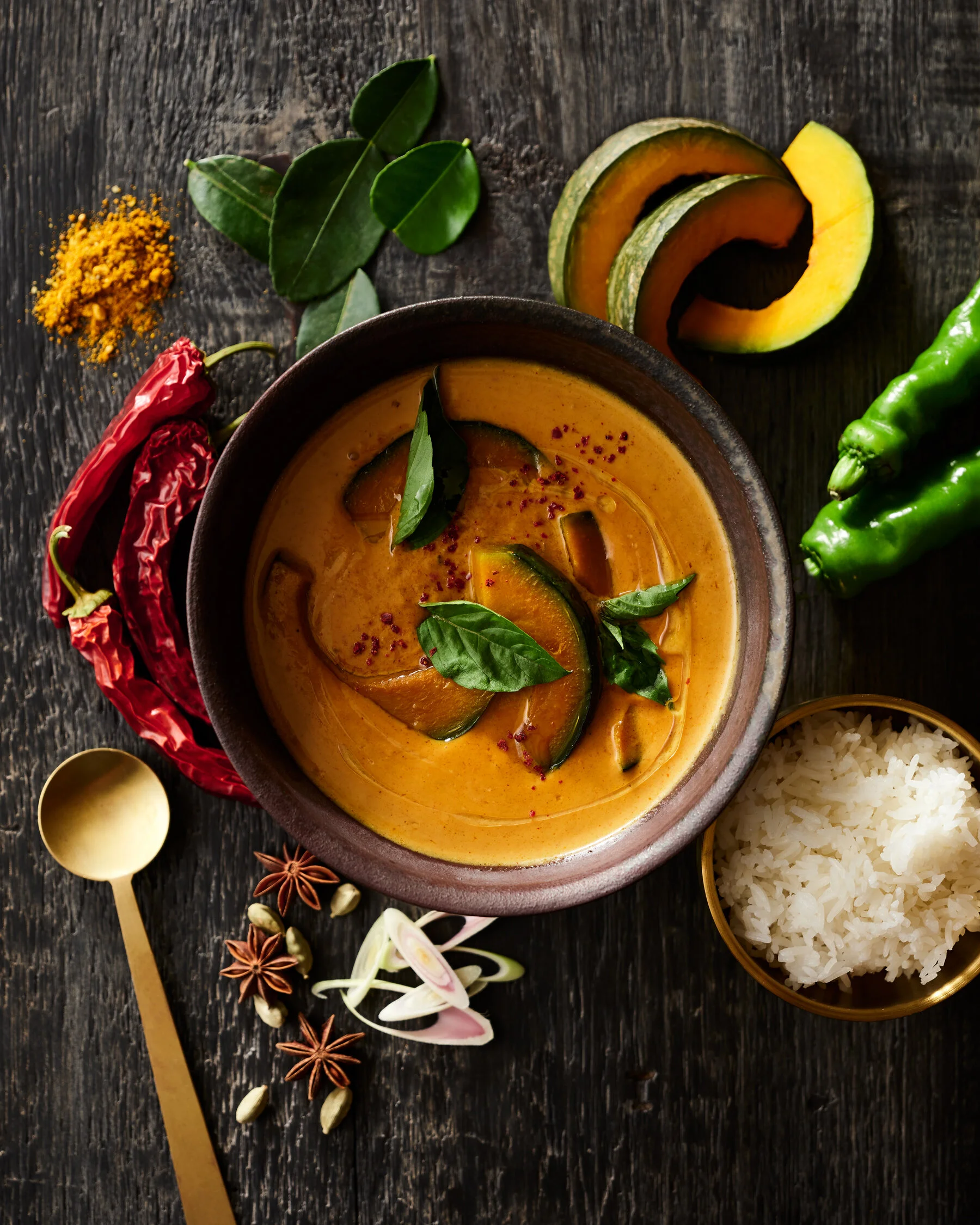 Bowl of pumpkin curry garnished with fresh basil leaves, surrounded by sliced pumpkin, green chili peppers, dried red chili peppers, star anise, ginger slices, cardamom pods, and a bowl of white rice on a dark wooden surface. Photographed for the coo