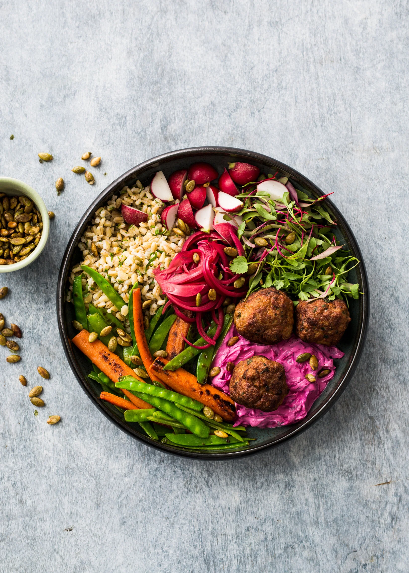 Vegan bowl with lentils, radishes, microgreens, shredded carrots, snap peas, falafel, beet hummus, and pumpkin seeds on a gray surface.