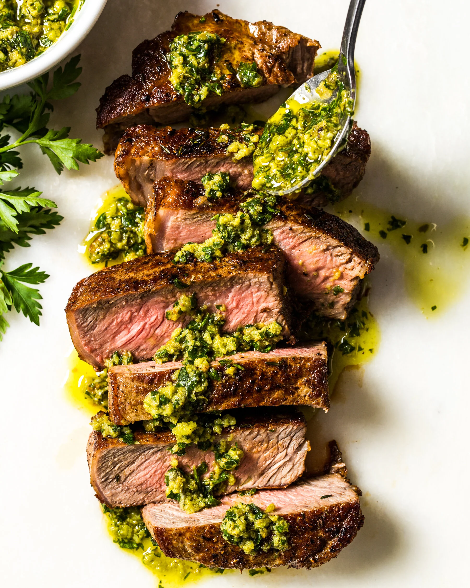Sliced medium-rare steak topped with green herb sauce on a white plate with parsley. Boston based food and hospitality photographer.