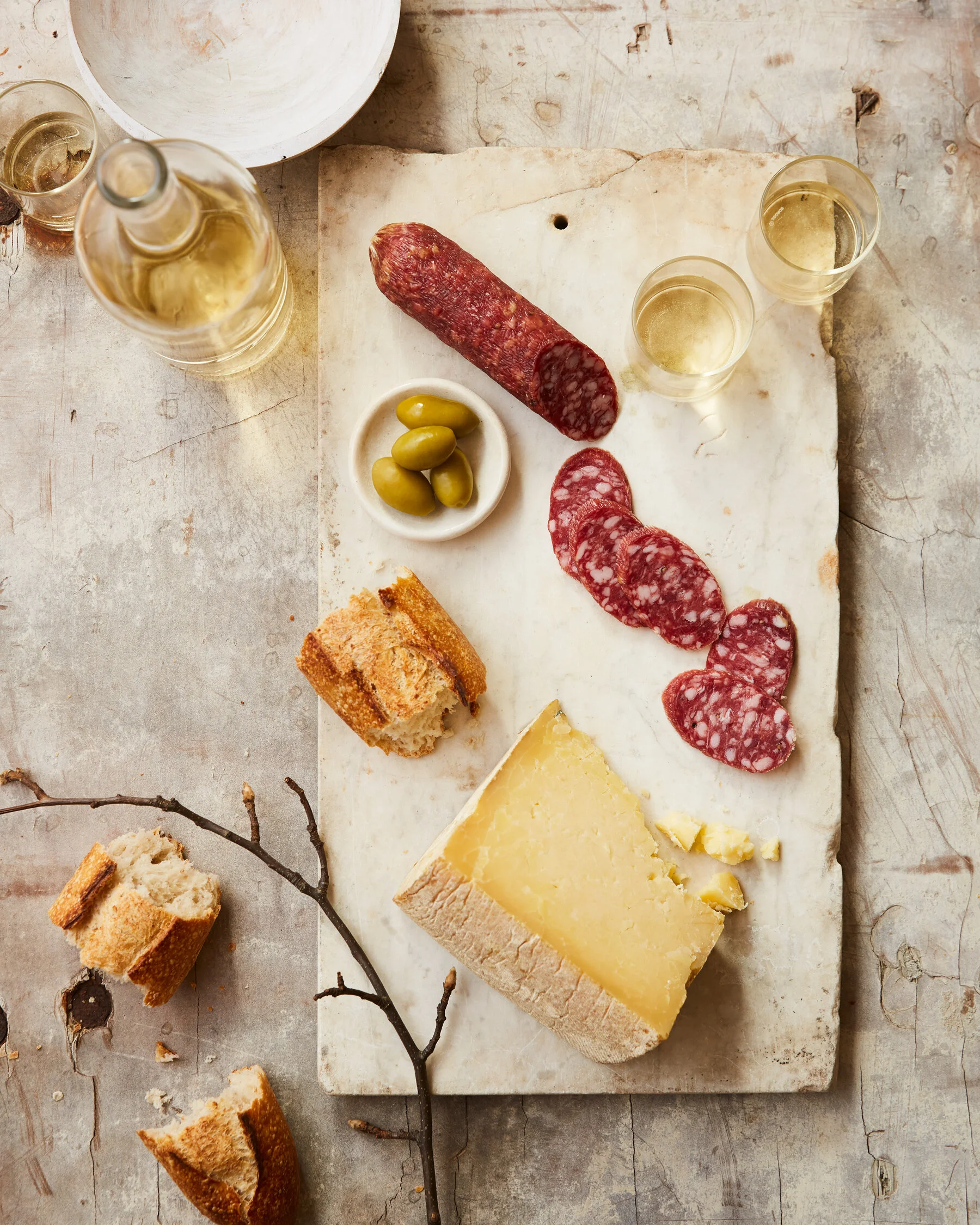 Charcuterie board with salami, cheese, green olives, bread, and glasses of white wine on a rustic table. Photographed for a cookbook about sausage making. Boston based food and hospitality photographer.