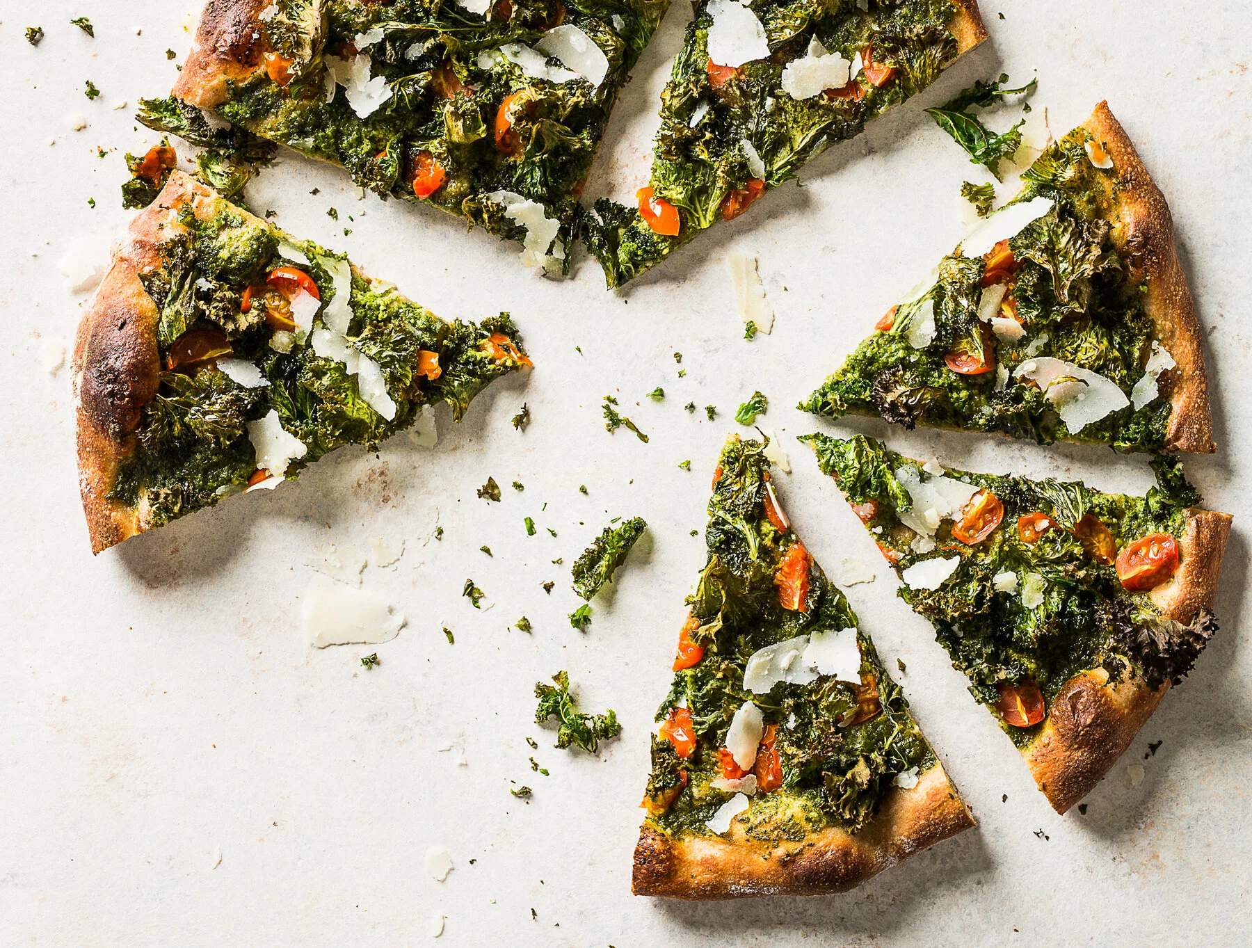 A sliced vegetable pizza with kale, cherry tomatoes, and shaved cheese on a white surface. Boston based food and hospitality photographer.