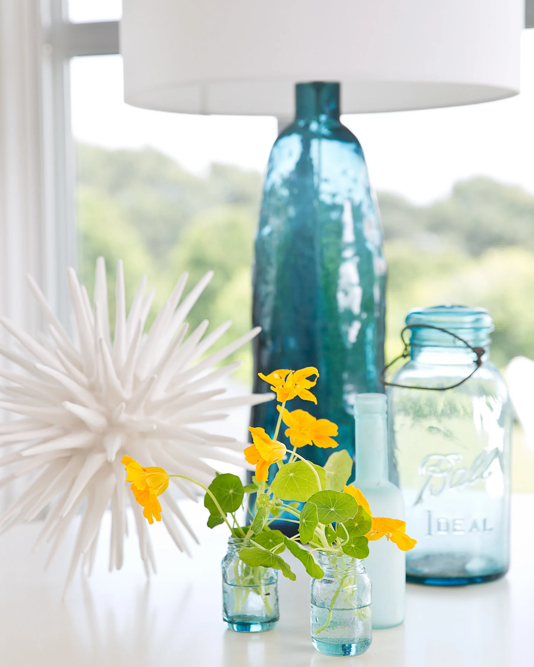 Decorative glass jars and vases with yellow flowers and white starburst sculpture, set on a white surface near a window.