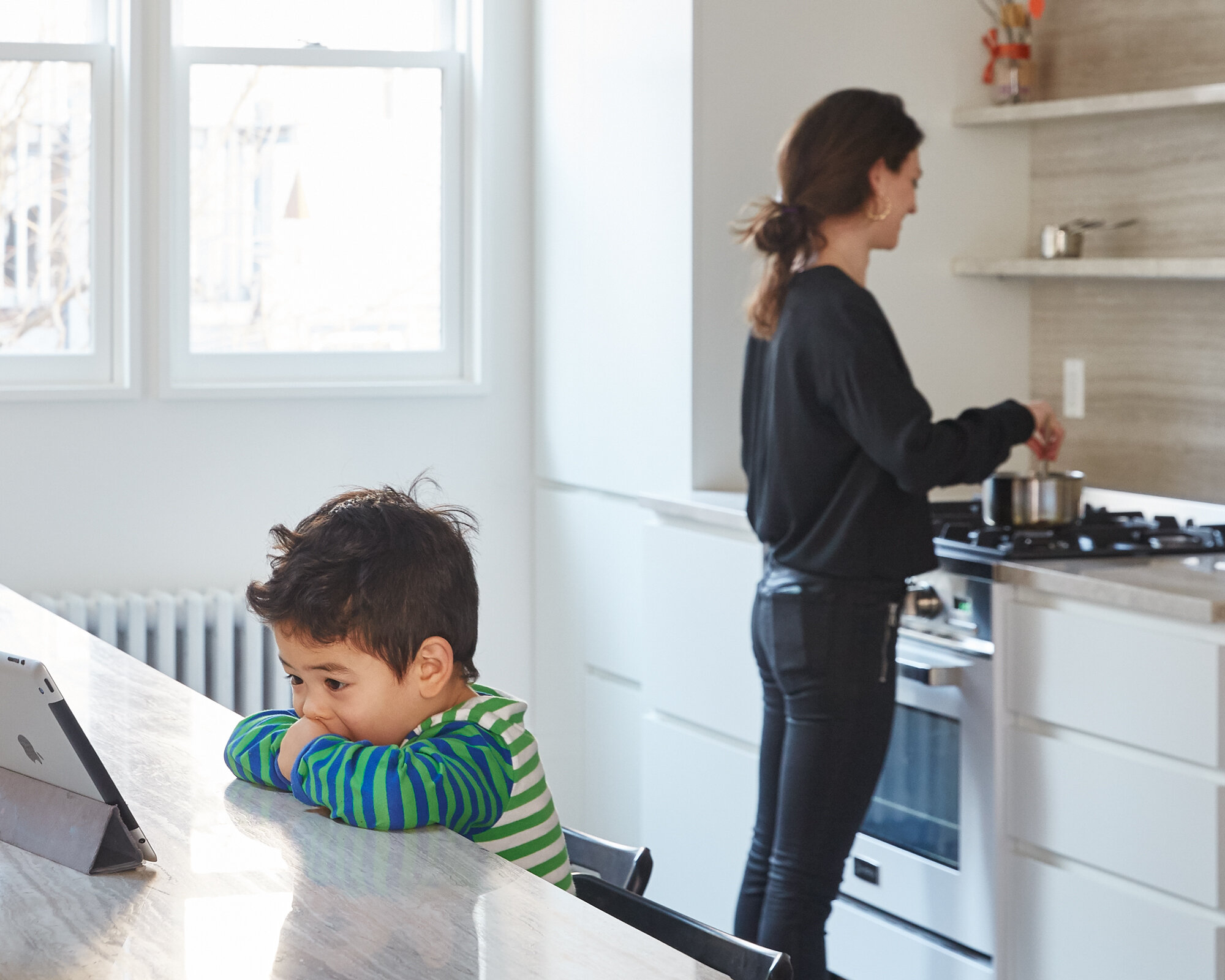 A young boy sitting at a kitchen table looking at a tablet, and a woman cooking on the stove in a bright kitchen with large windows.
