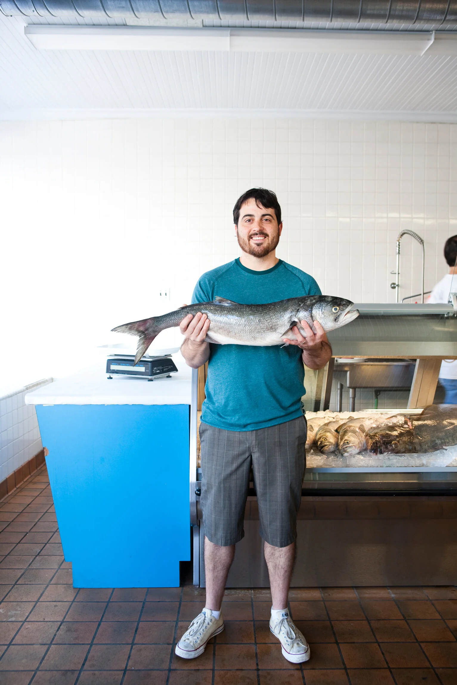 A man holding a large fish in a fish market or grocery store, smiling, with a background of tiled walls, a scale, and other fish on ice.