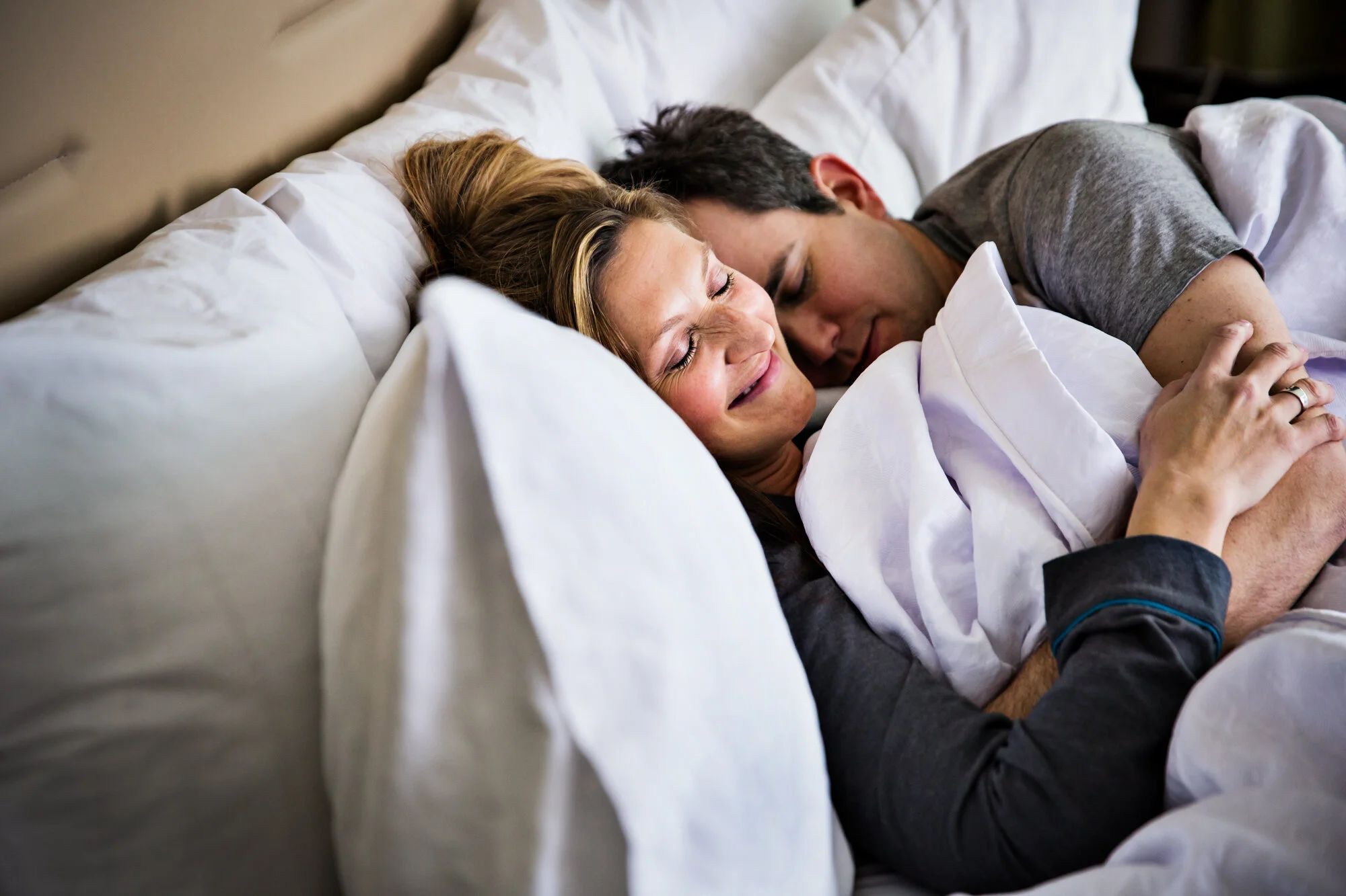 A happy couple is lying in bed, cuddling and smiling with their eyes closed.