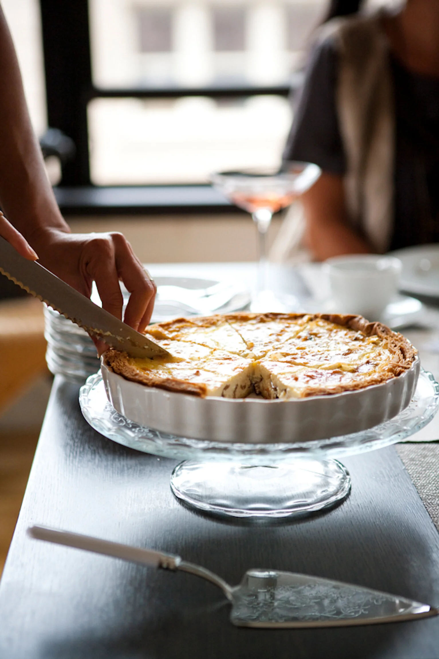 Person cutting a slice of baked cheese or potato dish on a glass cake stand at a dining table.