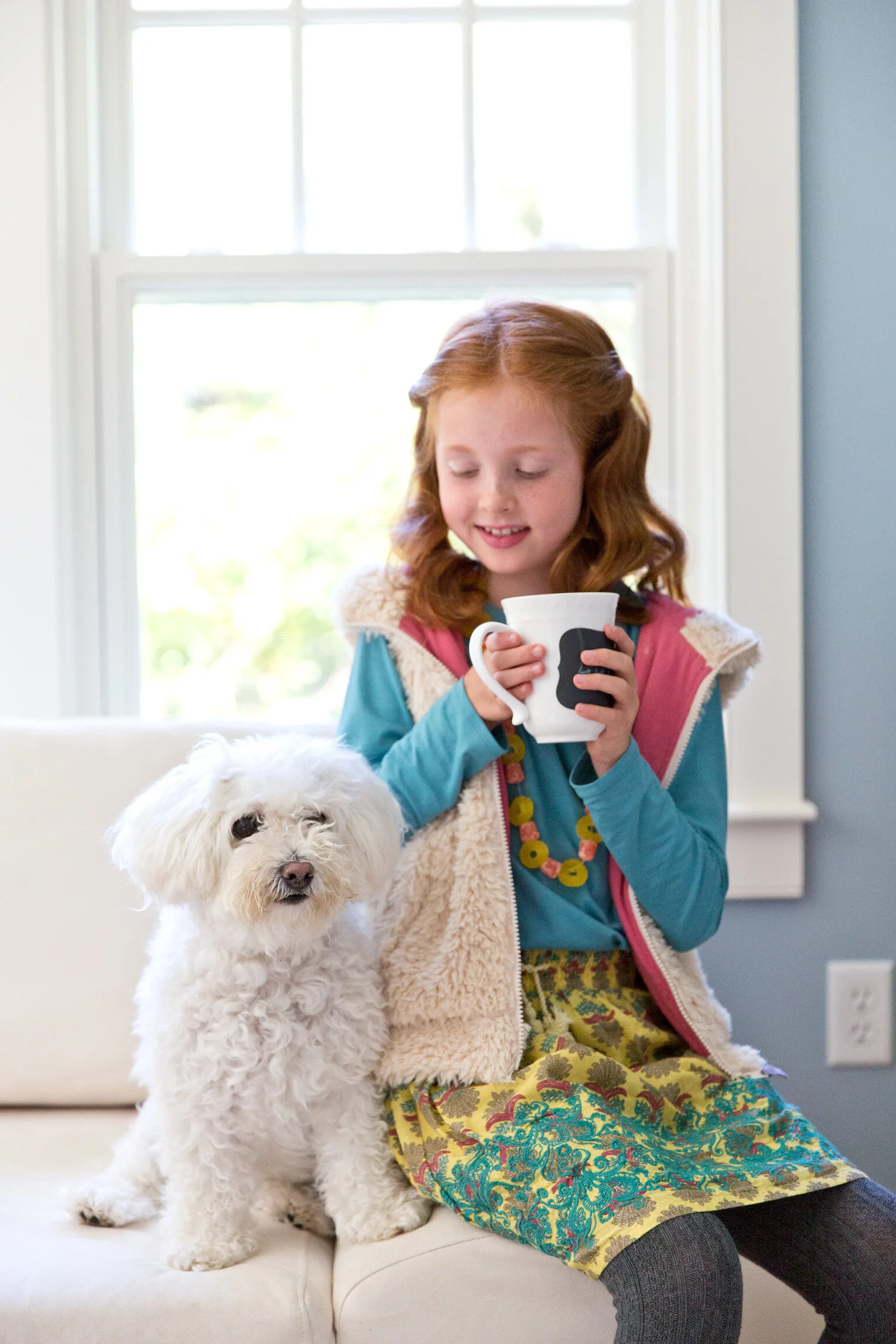 A young girl with red hair holding a white mug and sitting next to a fluffy white dog by a window