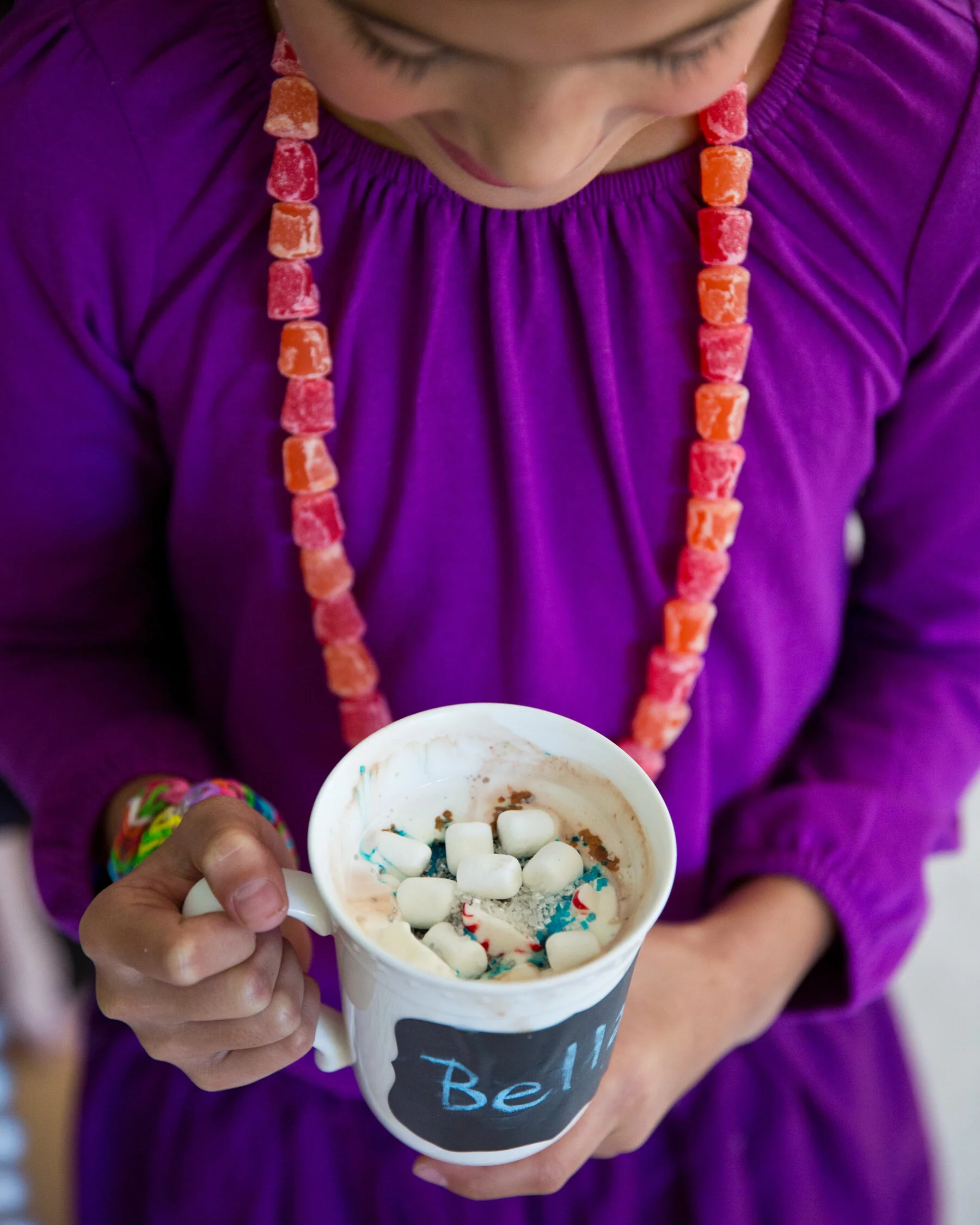 A girl in a purple shirt wearing a beaded necklace holding a mug filled with hot chocolate topped with mini marshmallows and colorful sprinkles.