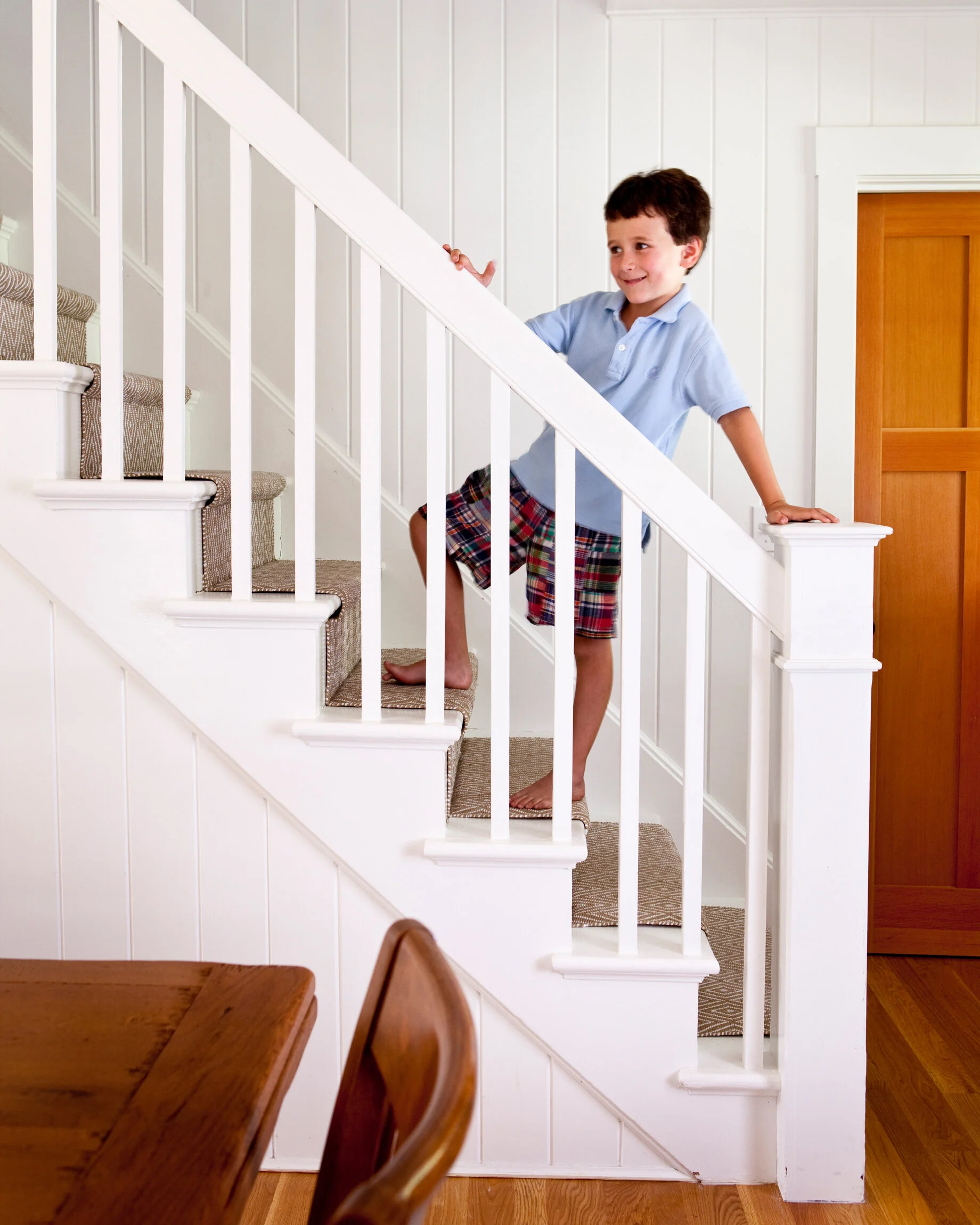 A young boy with short dark hair, wearing a light blue polo shirt and plaid shorts, is climbing a staircase inside a home. He is smiling and holding onto the railing with one hand, while stepping on the carpeted stairs with one foot and balancing wit