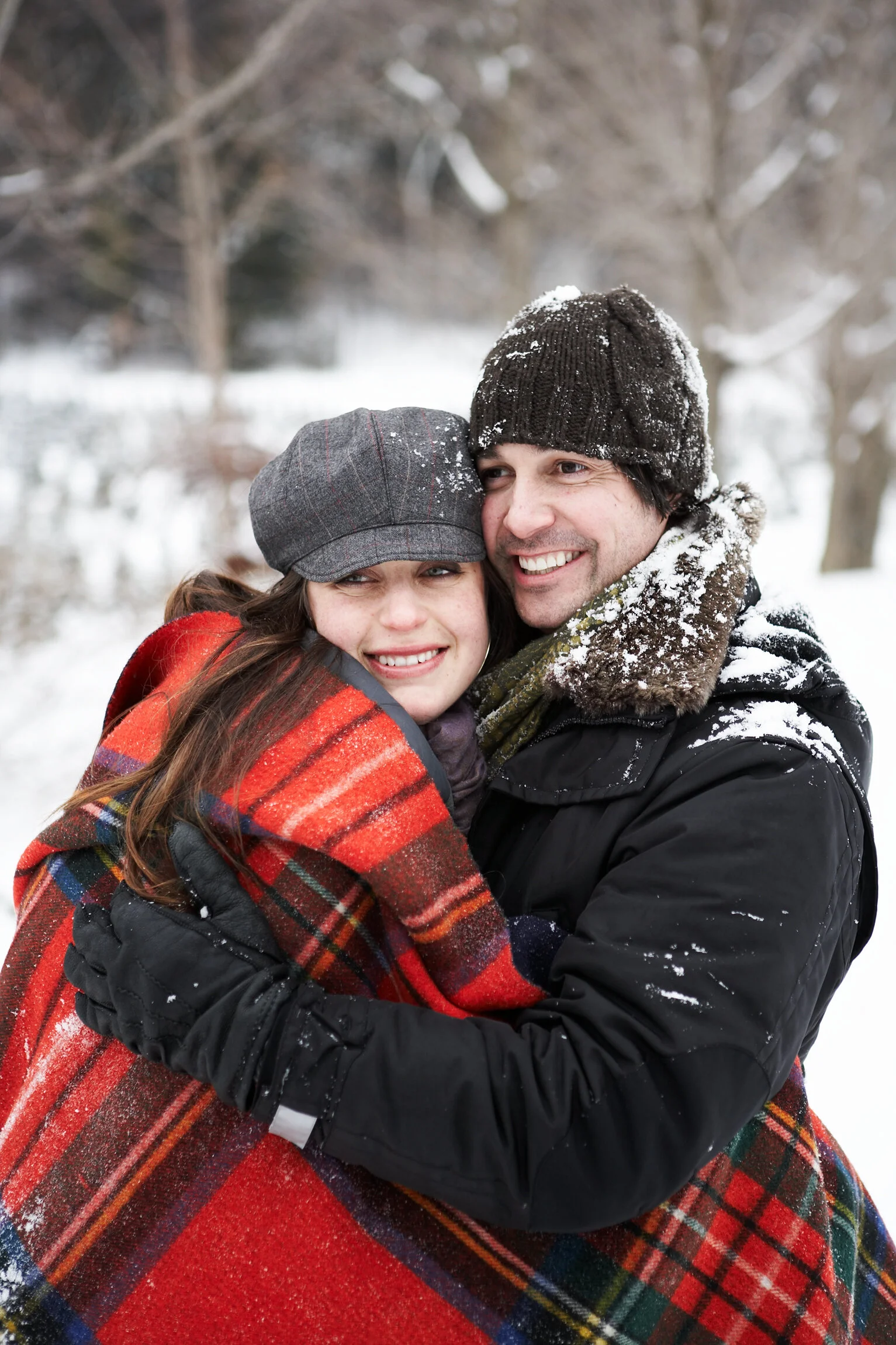 A smiling couple in winter clothing embraces outdoors in a snowy landscape.