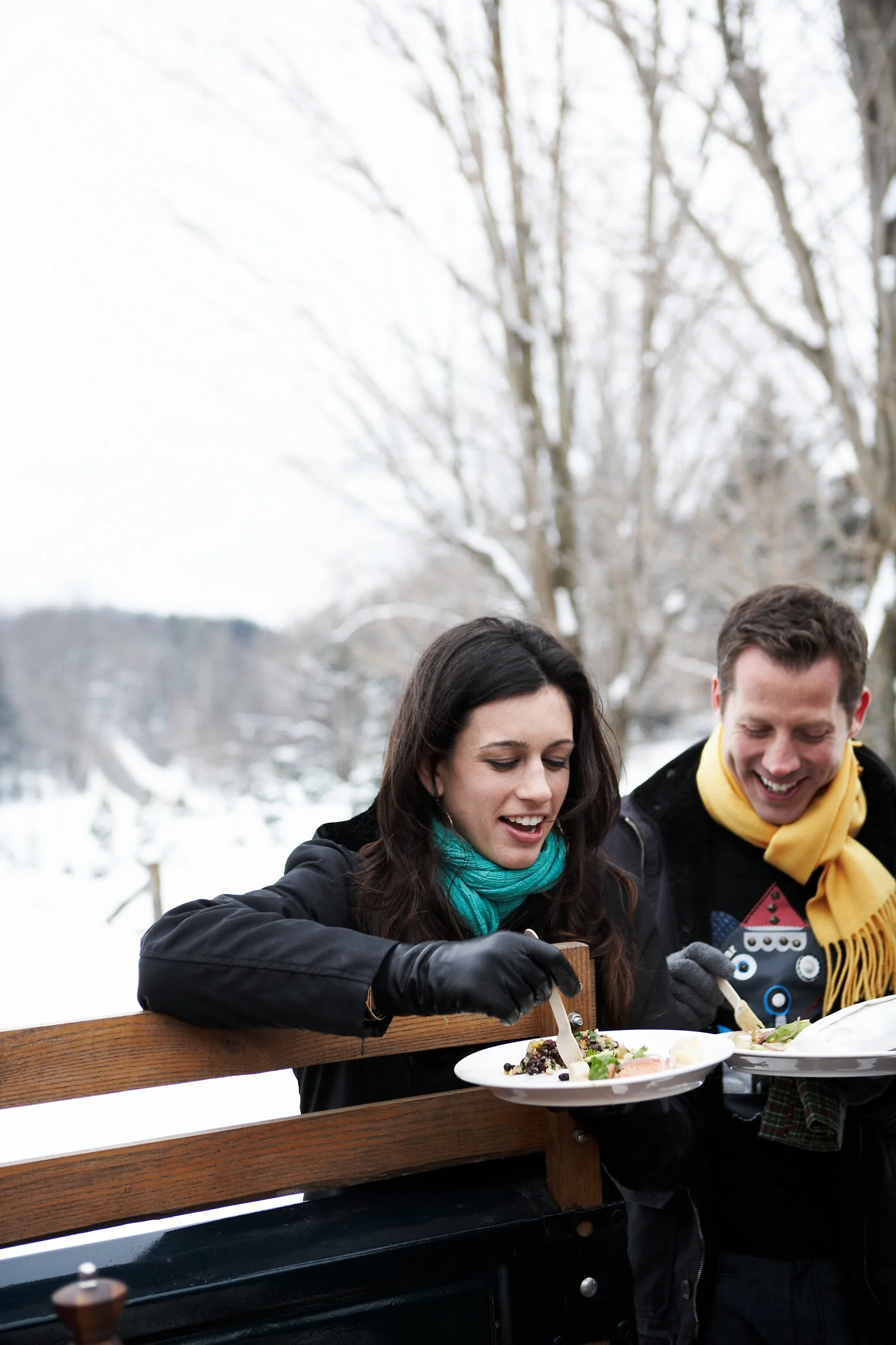 A woman and a man sitting on a park bench outdoors in winter, smiling and eating food from plates.