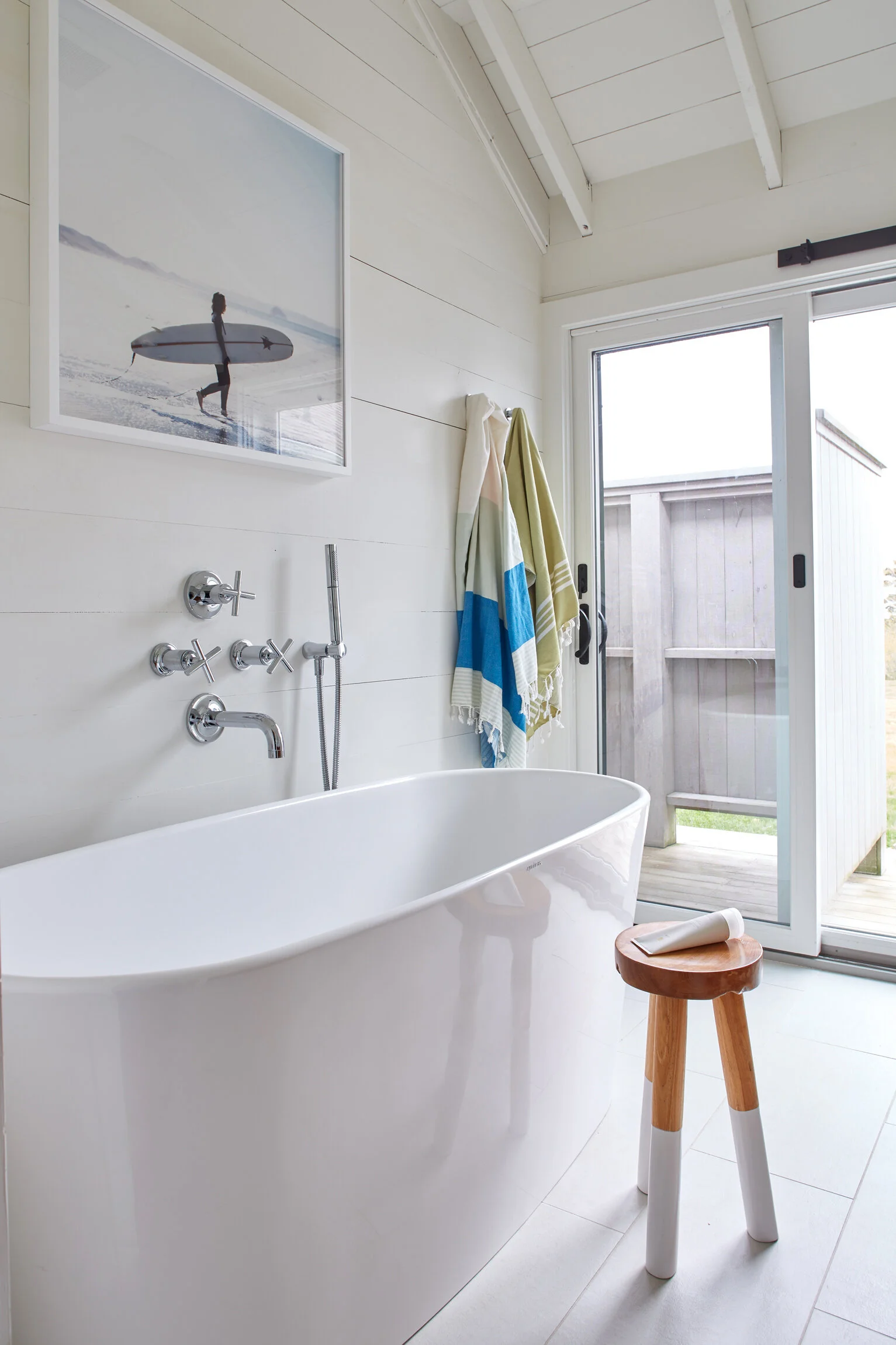 Modern bathroom with a freestanding bathtub, wooden stool, wall-mounted faucet, and sliding glass door leading outside