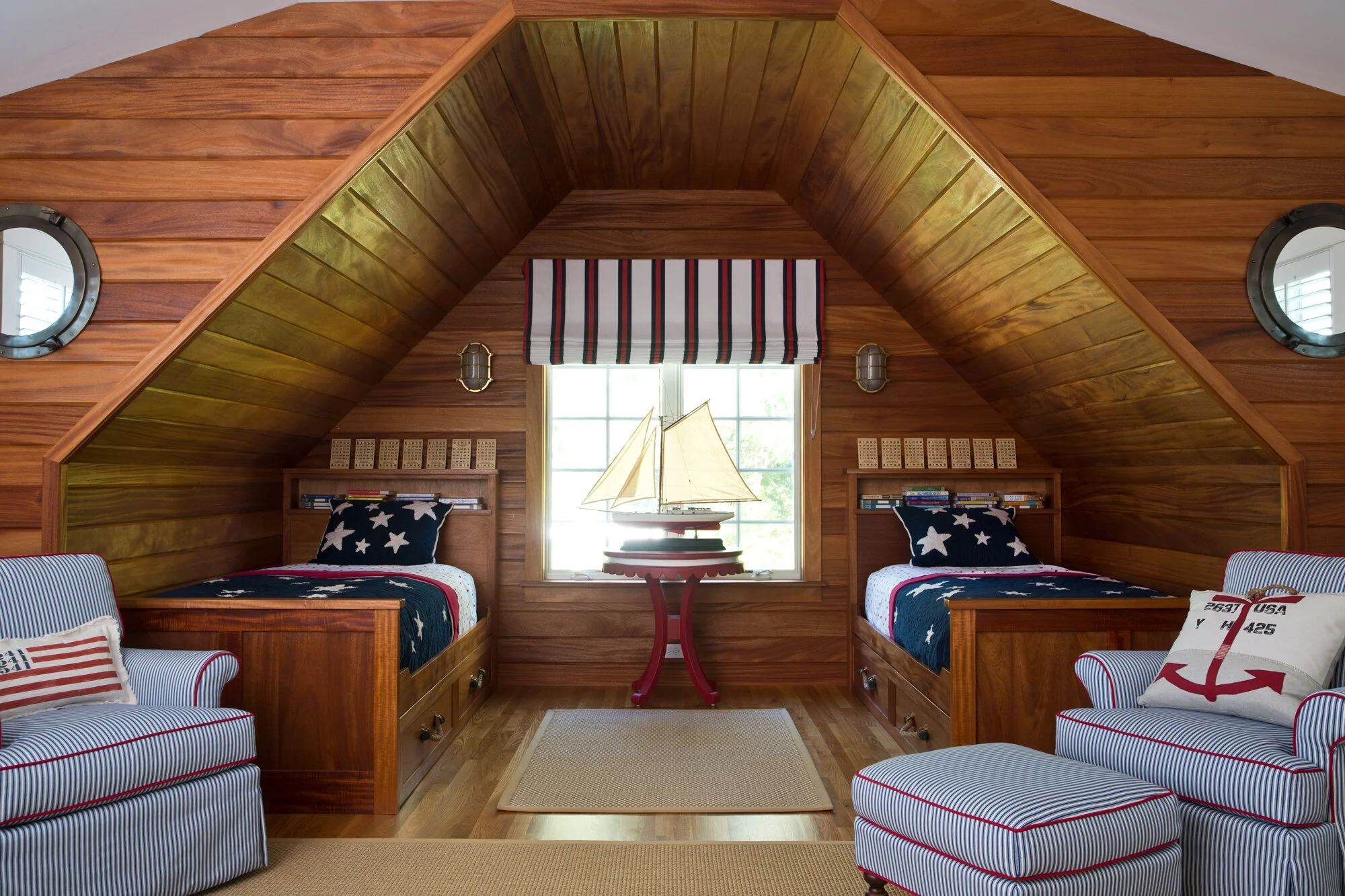 A nautical-themed bedroom with two wooden beds with star and stripe bedding, striped cushions, striped valance, a table with a model sailboat, and Porthole-style windows.