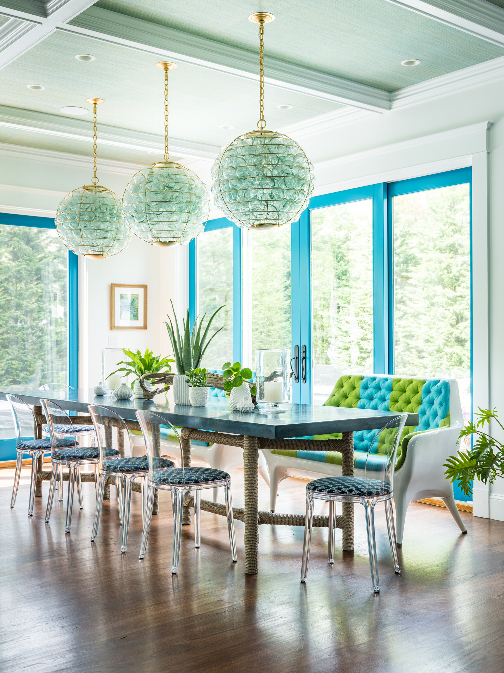 Bright dining room with large windows, modern chandelier with glass orbs, and a mix of colorful and clear seating around a table with plants.