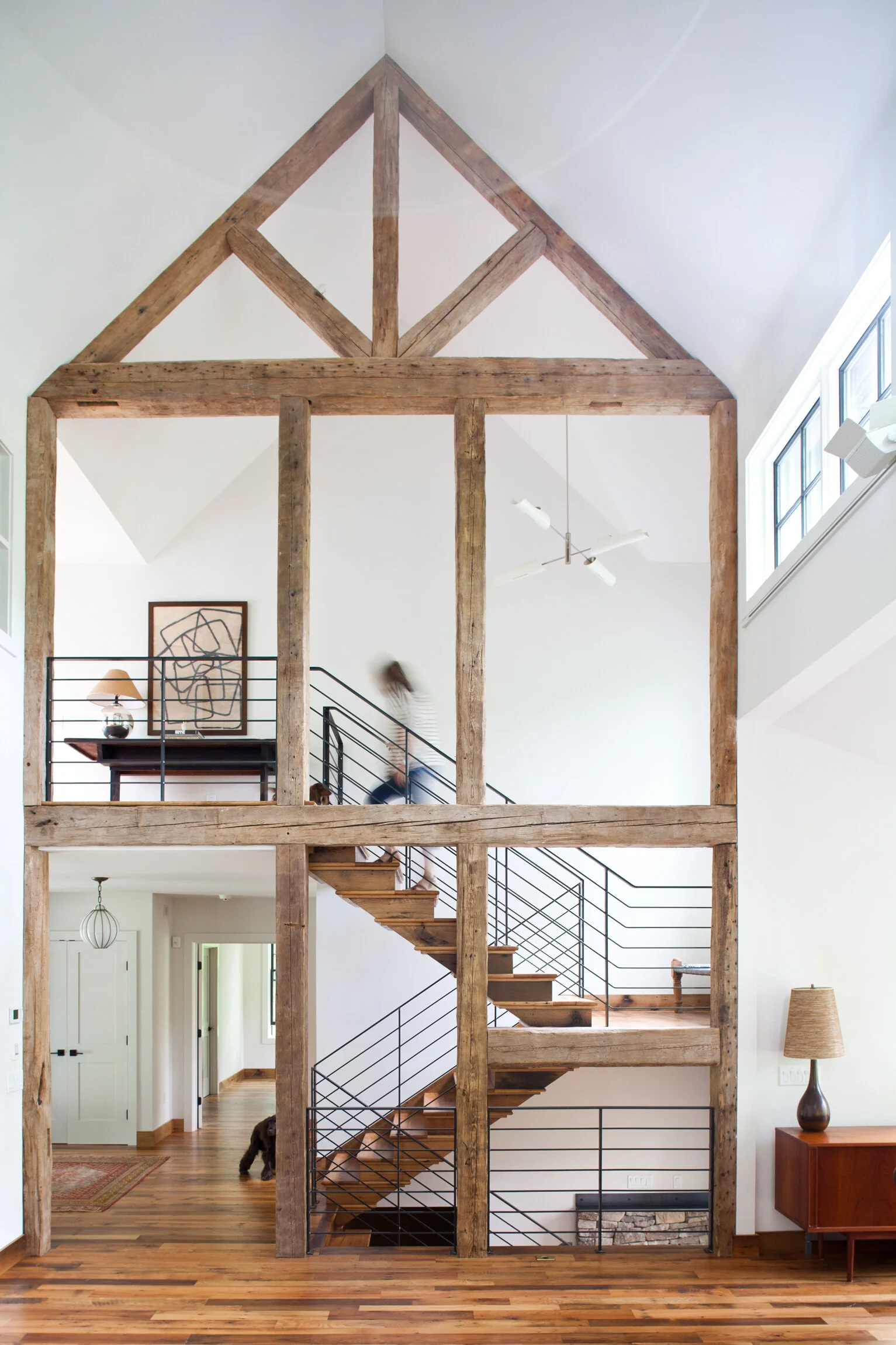Interior view of a modern home with a prominent wooden staircase and exposed wooden beams, with a person walking upstairs.