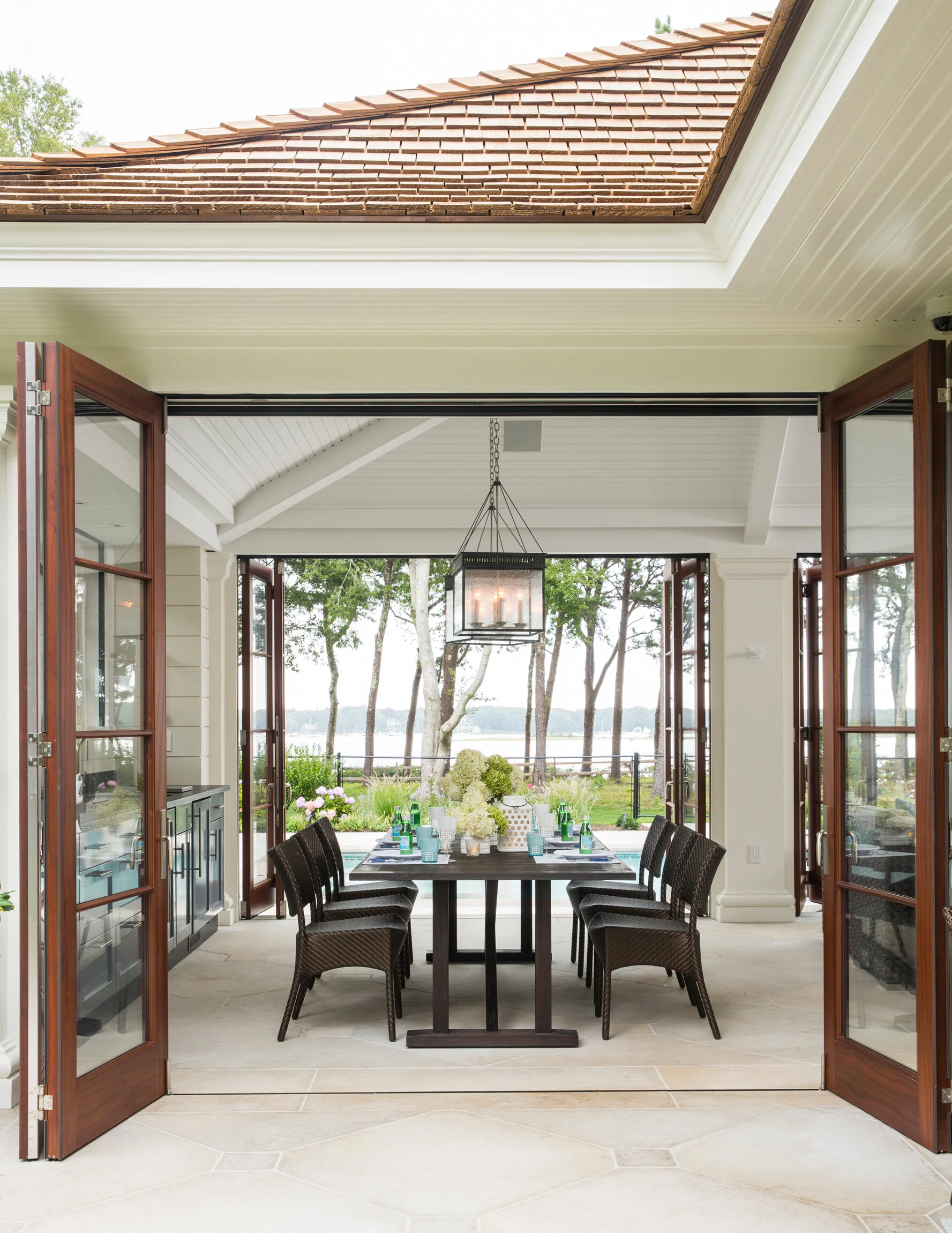 Dining area outdoors with a black table, wicker chairs, and a view of trees and water through open glass doors.