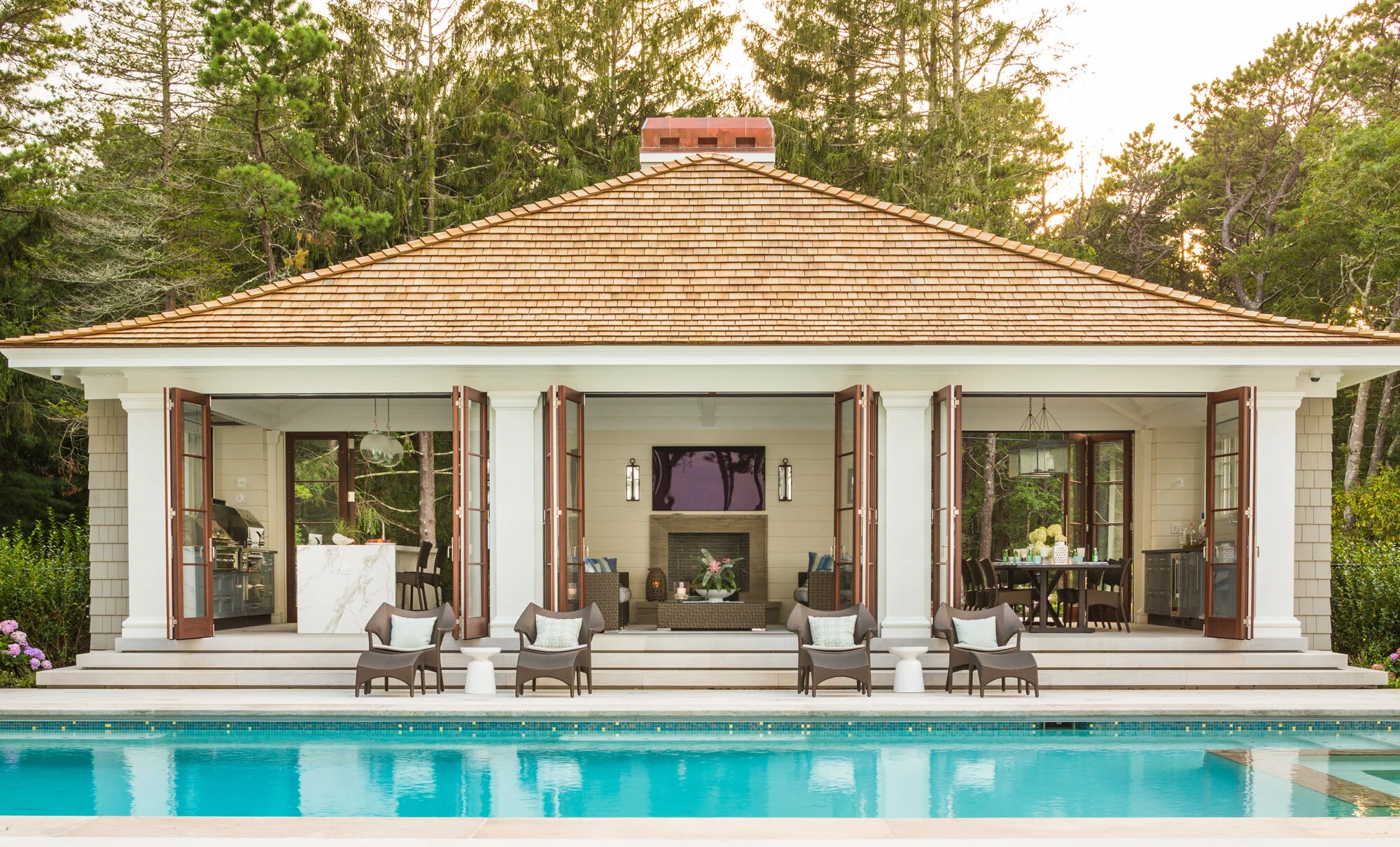 A backyard with a swimming pool in front and a house with an outdoor living area in the background. The house has large open folding doors, a tiled roof, and a fireplace with a TV mounted above. There are chairs and tables on the patio, and trees sur