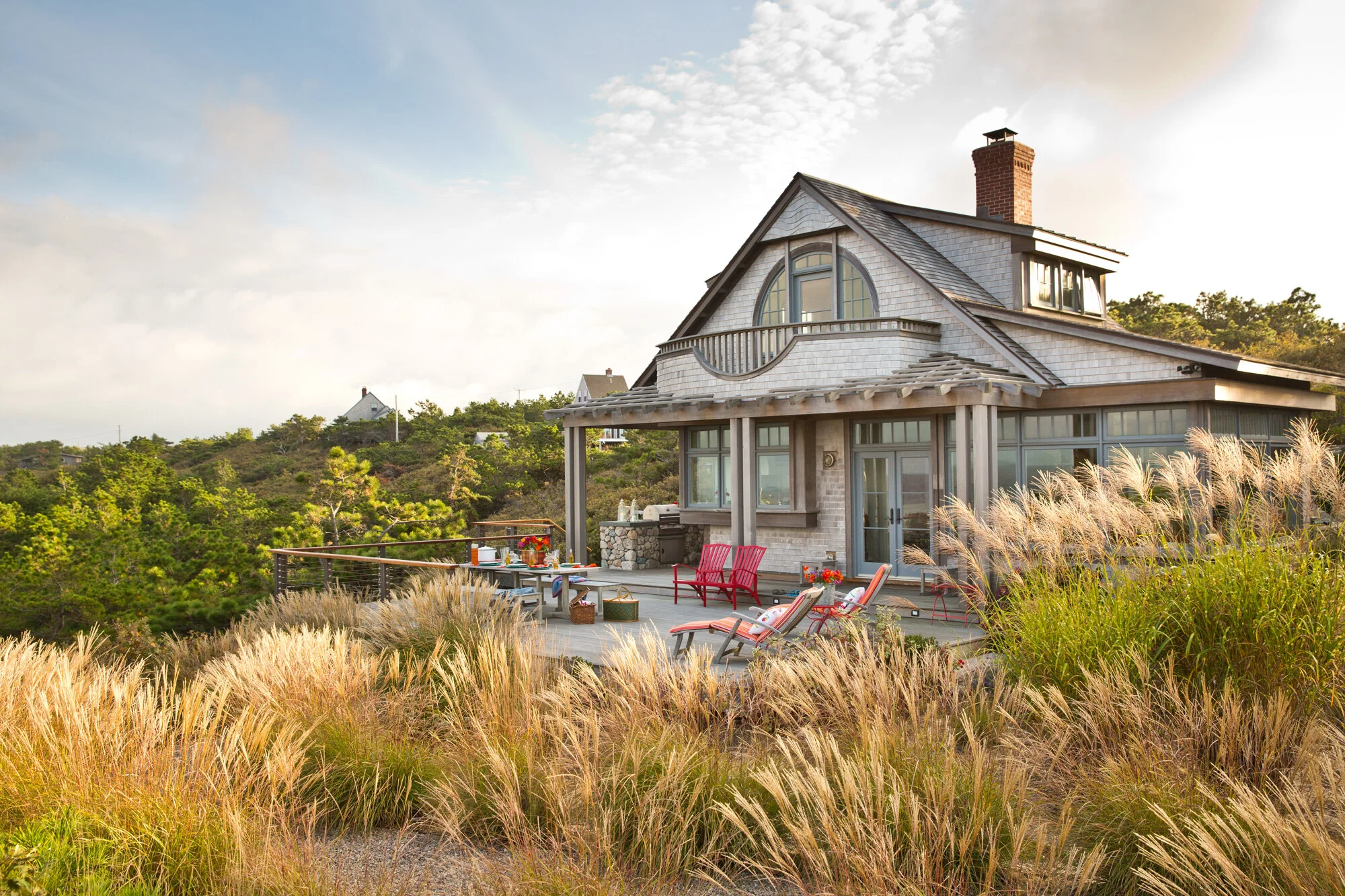 A house on a hill with a large outdoor deck decorated with chairs, a table, and outdoor accessories, surrounded by tall grasses, with trees and other houses in the background under a partly cloudy sky.