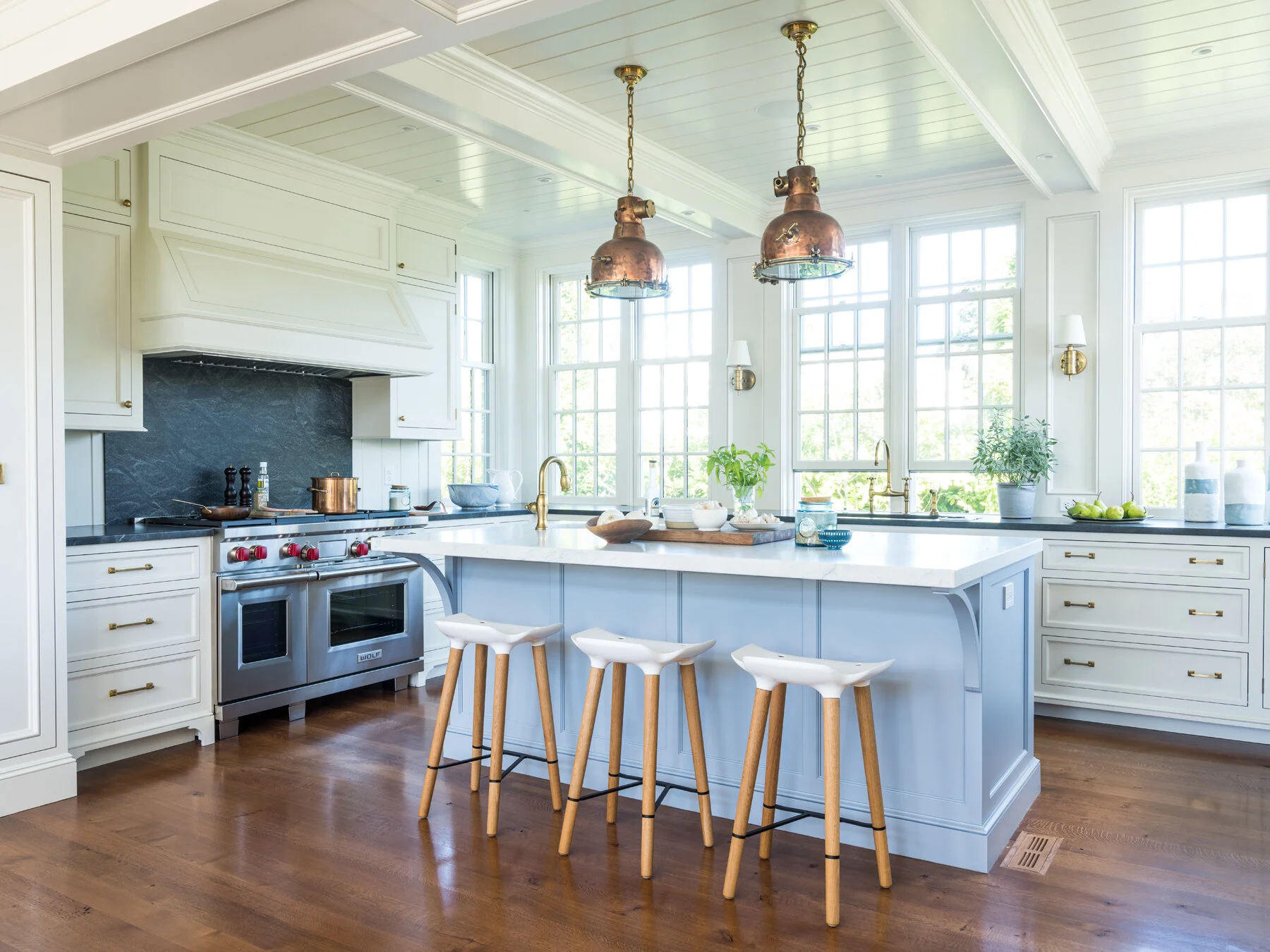 Bright kitchen with white cabinets, a large island with three white and wood stools, three copper pendant lights, large windows, and wooden floors.