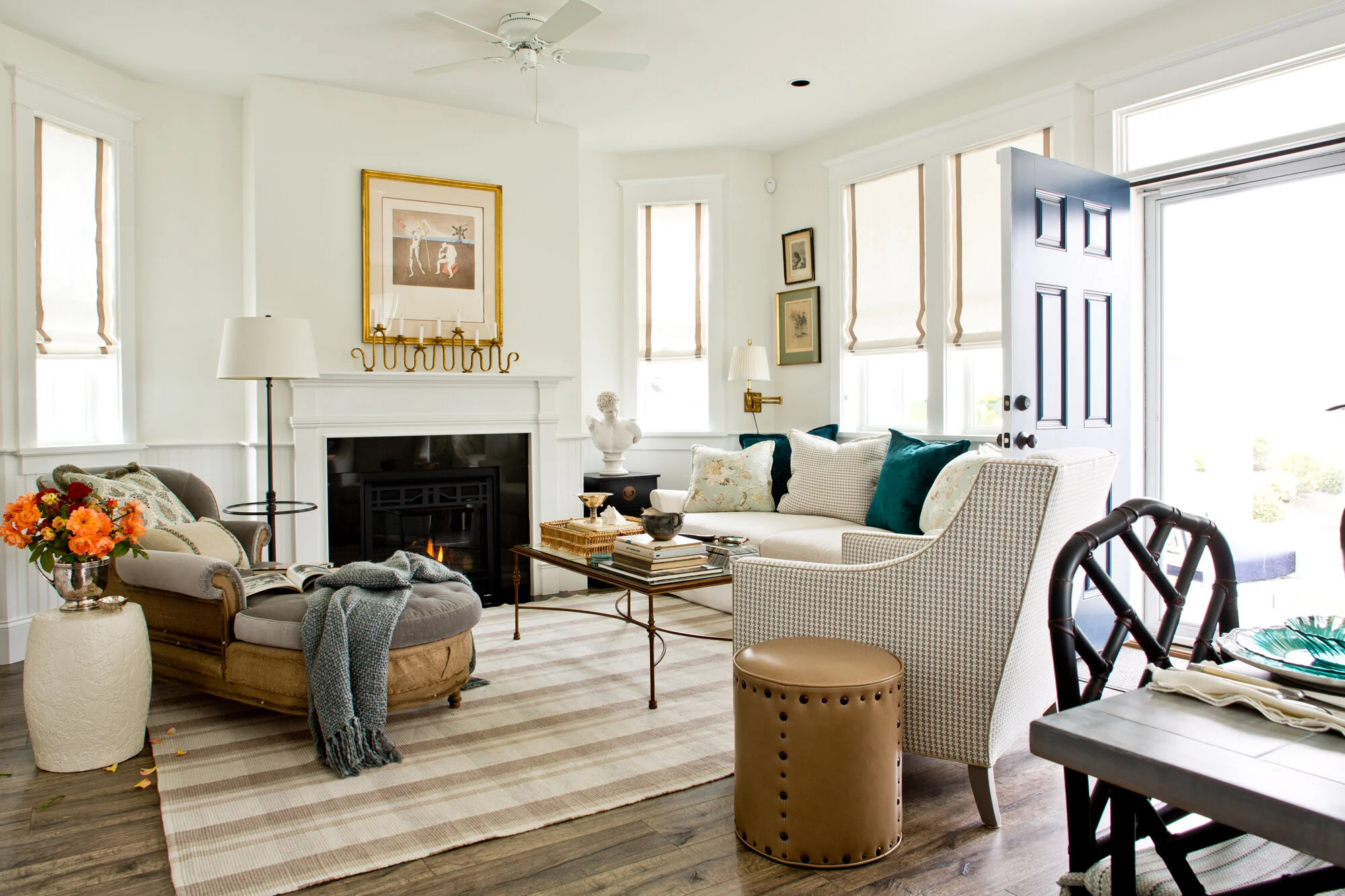 Bright living room with white walls, a fireplace, various seating including a beige armchair and a white sofa, a wooden coffee table with books and decor, and large windows letting in natural light.