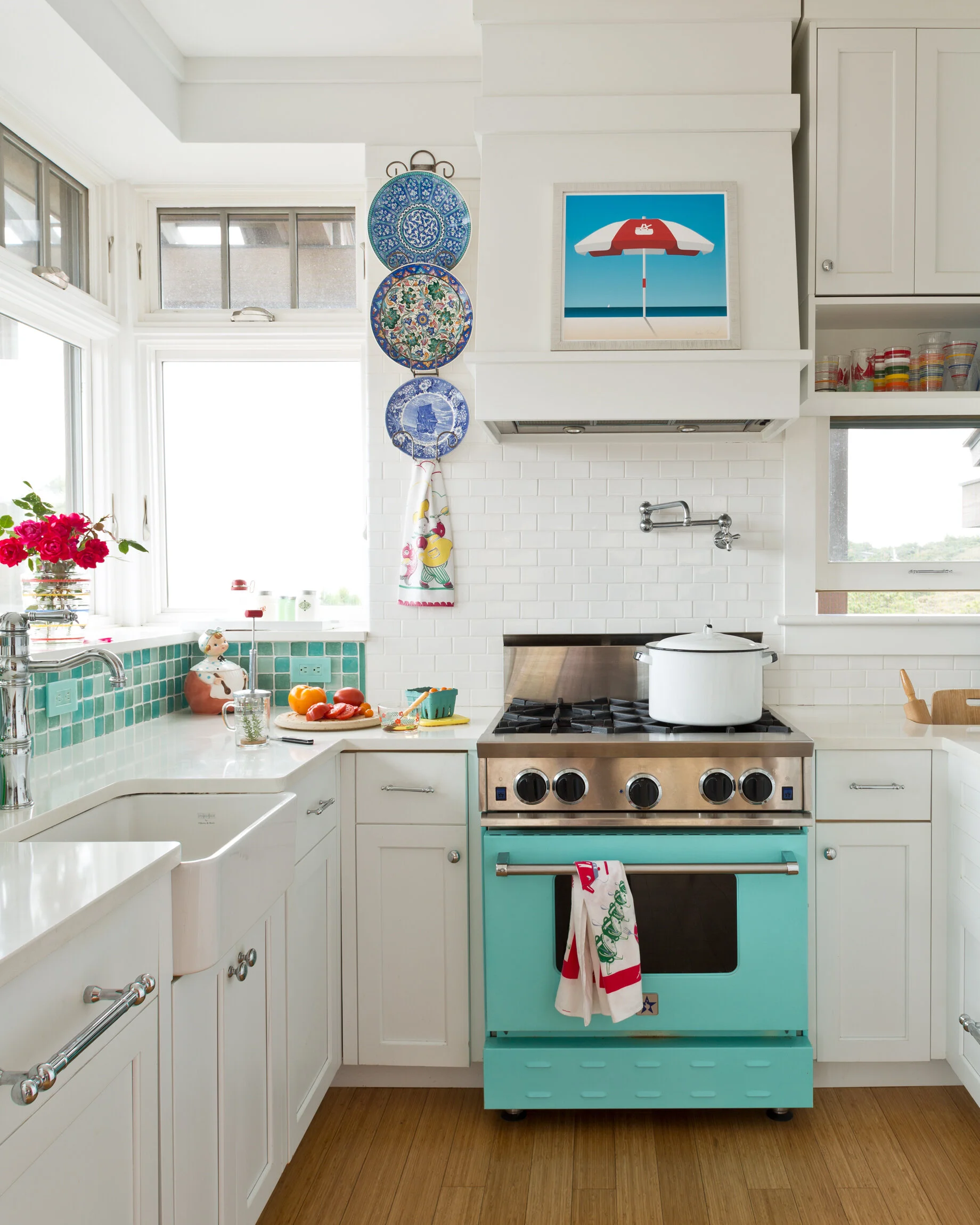 Bright kitchen with white cabinets, a teal stove, a white sink, and a window with pink flowers on the sill. There are plates hanging on the wall, a framed beach umbrella picture, and a countertop with vegetables and cooking utensils.