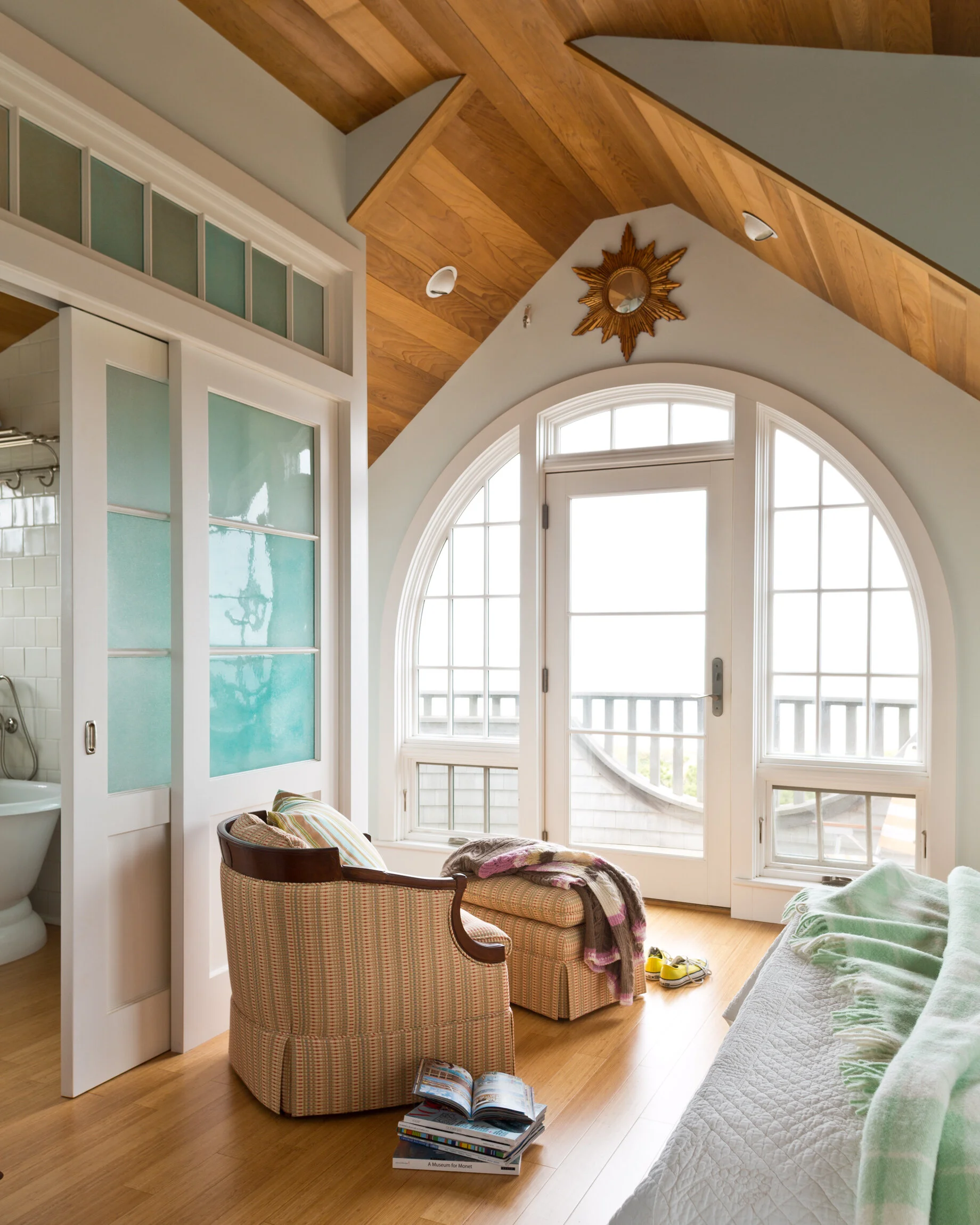 Bright living room with large arched window, wooden ceiling, patterned armchair with striped pillow, quilted sofa with green blanket, small ottoman with a throw blanket, open books on the floor, and a view of a balcony outside.