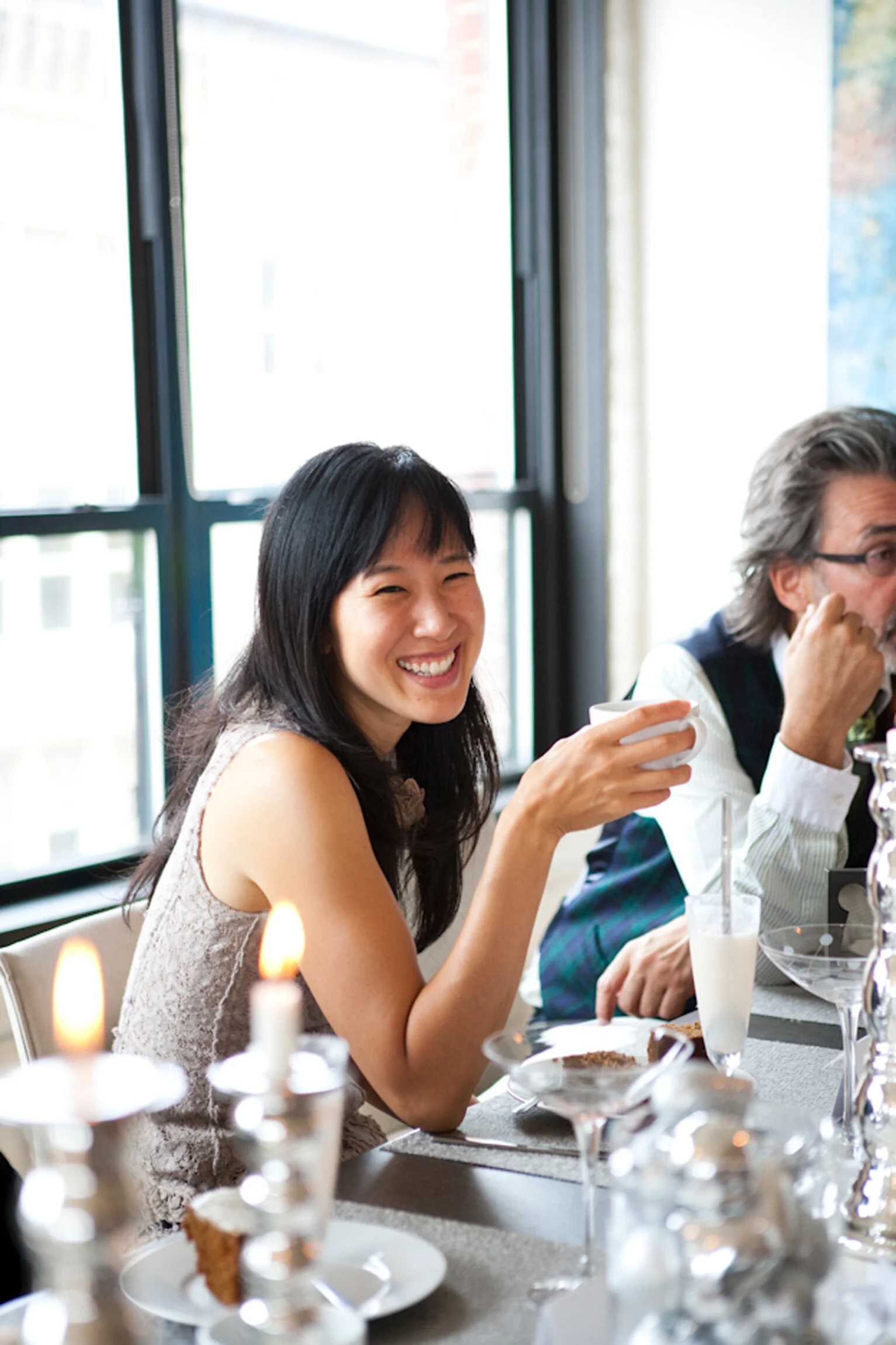 A woman smiling and holding a white cup at a table with food and drinks, with large windows in the background. Boston based food and hospitality photographer. Lifestyle photography.