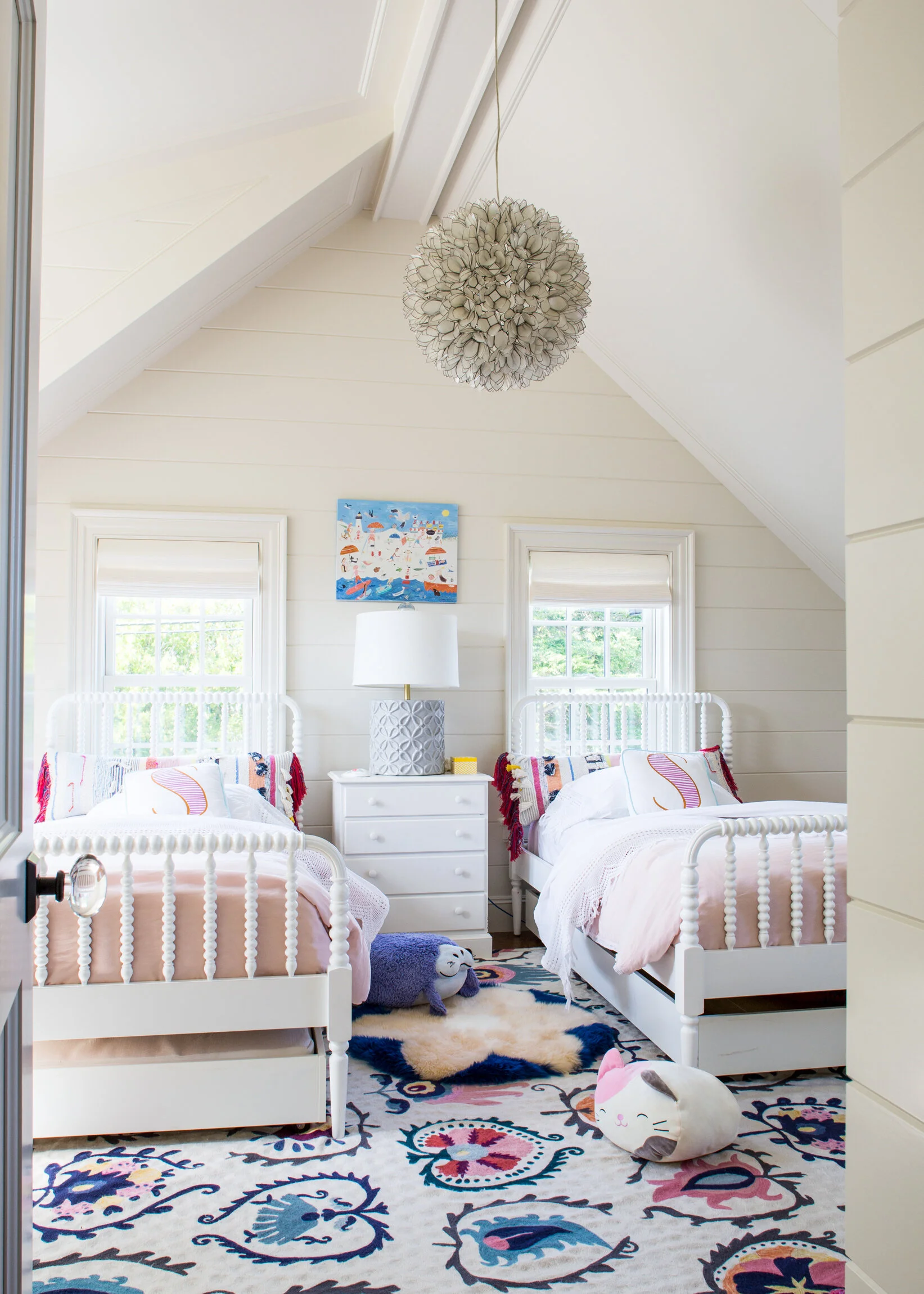 A bedroom with two white twin beds, a white dresser with a lamp, two windows, and decorative stuffed animals on the floor. The room has white wooden walls and a colorful patterned rug. Boston based interiors and architectural photographer.