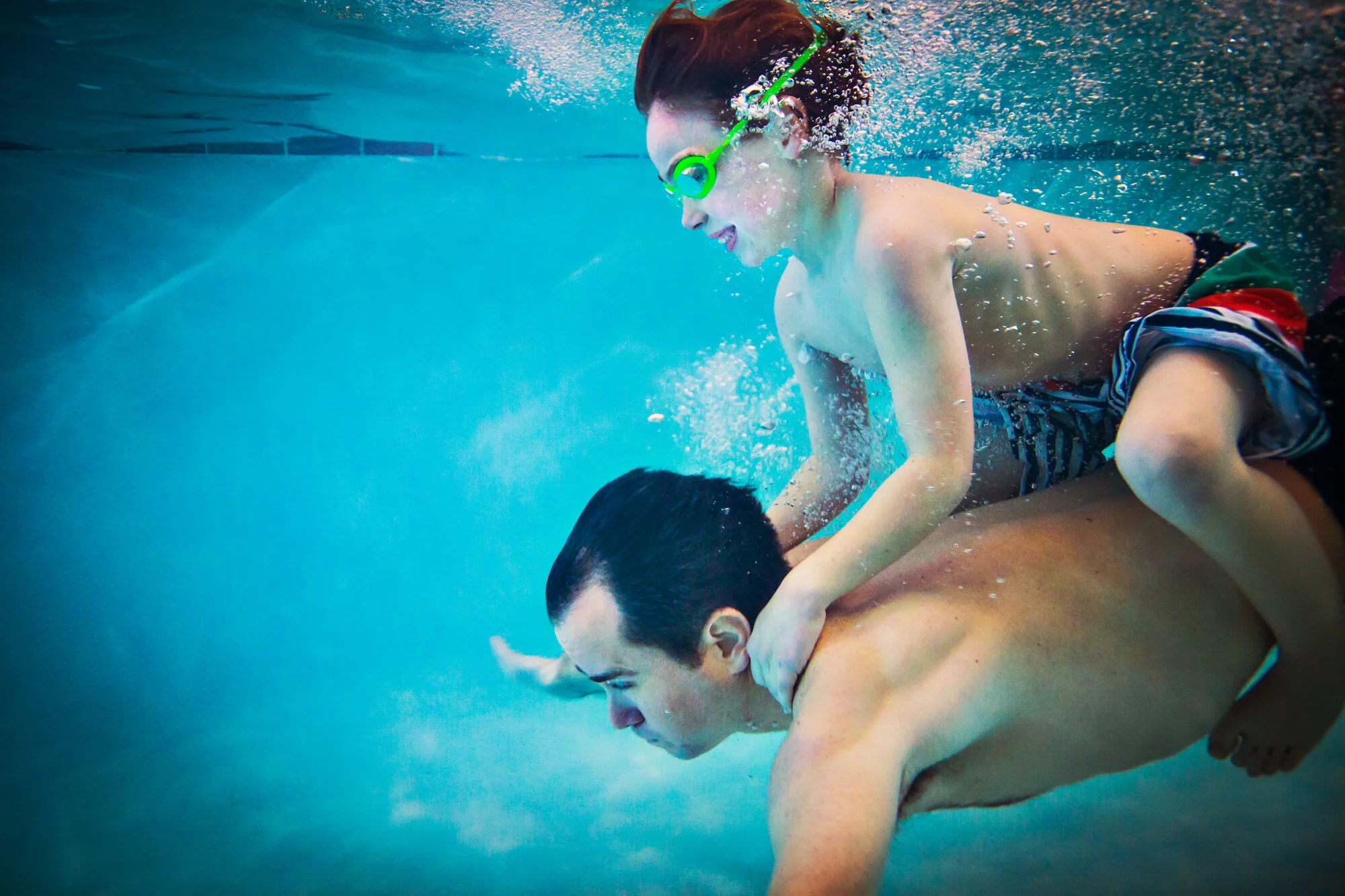 A young boy with goggles riding on the back of a man underwater in a swimming pool. Boston lifestyle and hospitality photographer.