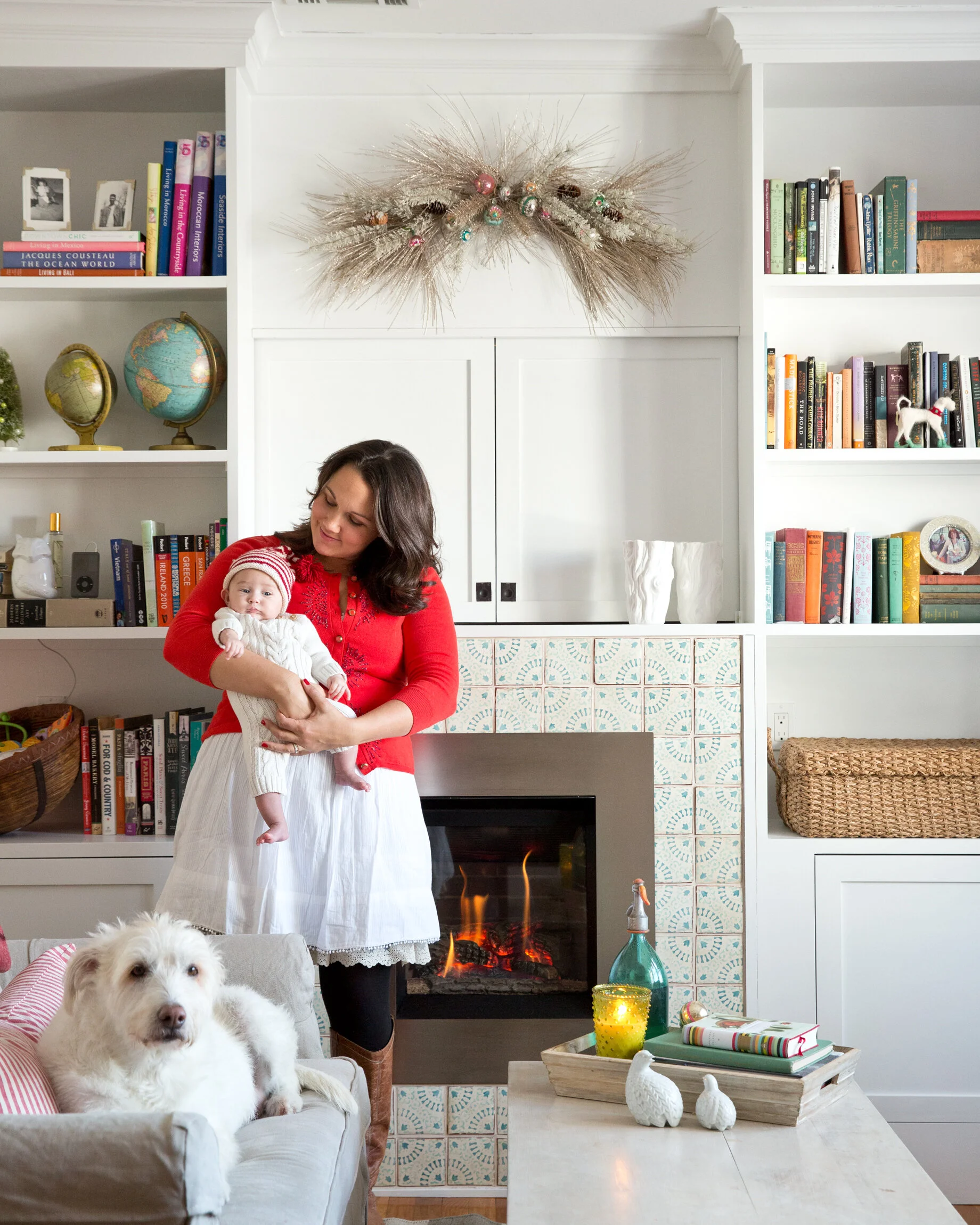A woman in a red sweater holding a baby in front of a fireplace decorated with holiday ornaments and a snowy white and beige interior with bookshelves, globe, and holiday decor. Boston lifestyle and hospitality photographer.