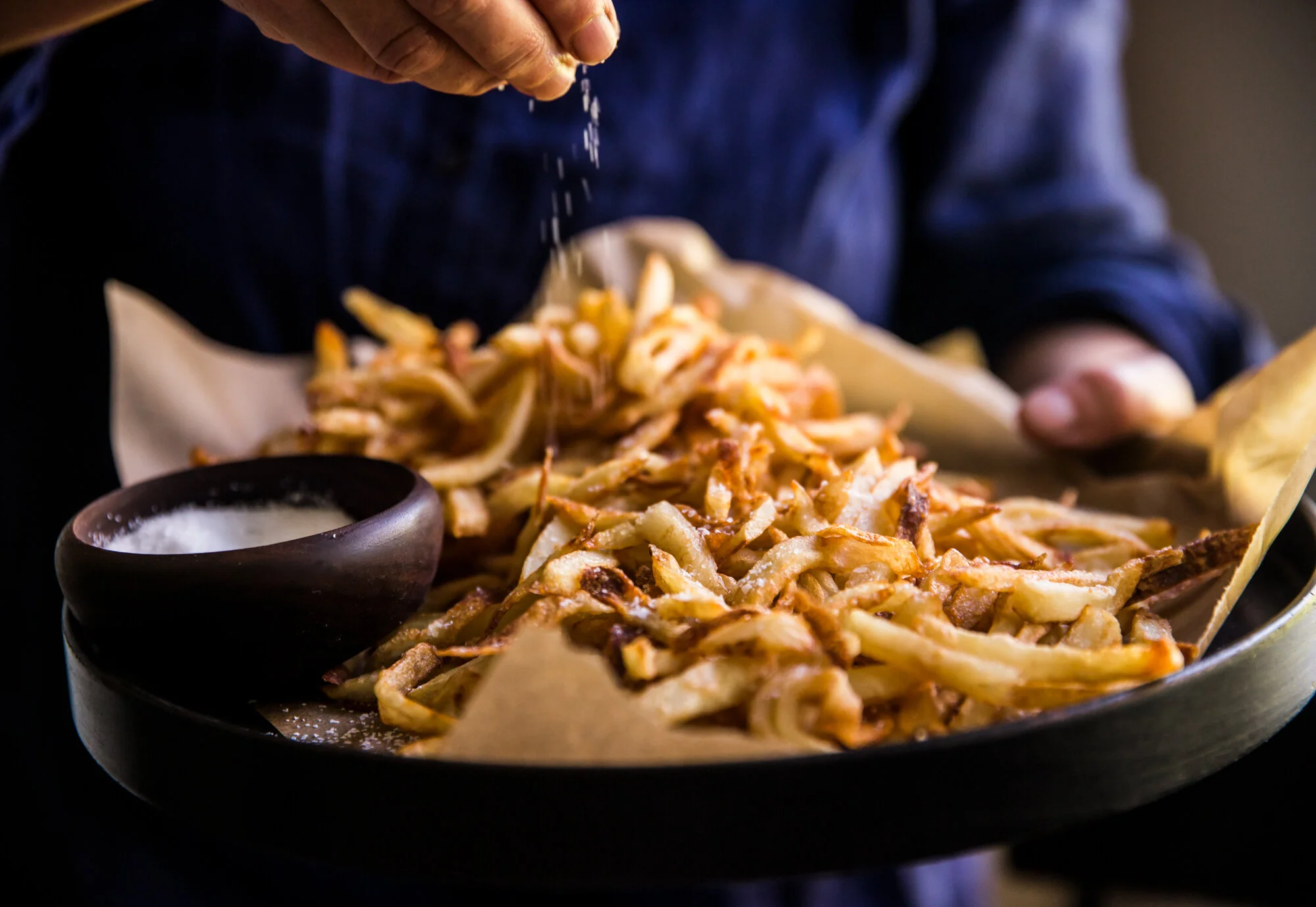 Person sprinkling salt onto a tray of cooked French fries with a small bowl of salt nearby. Boston based food and hospitality photographer.