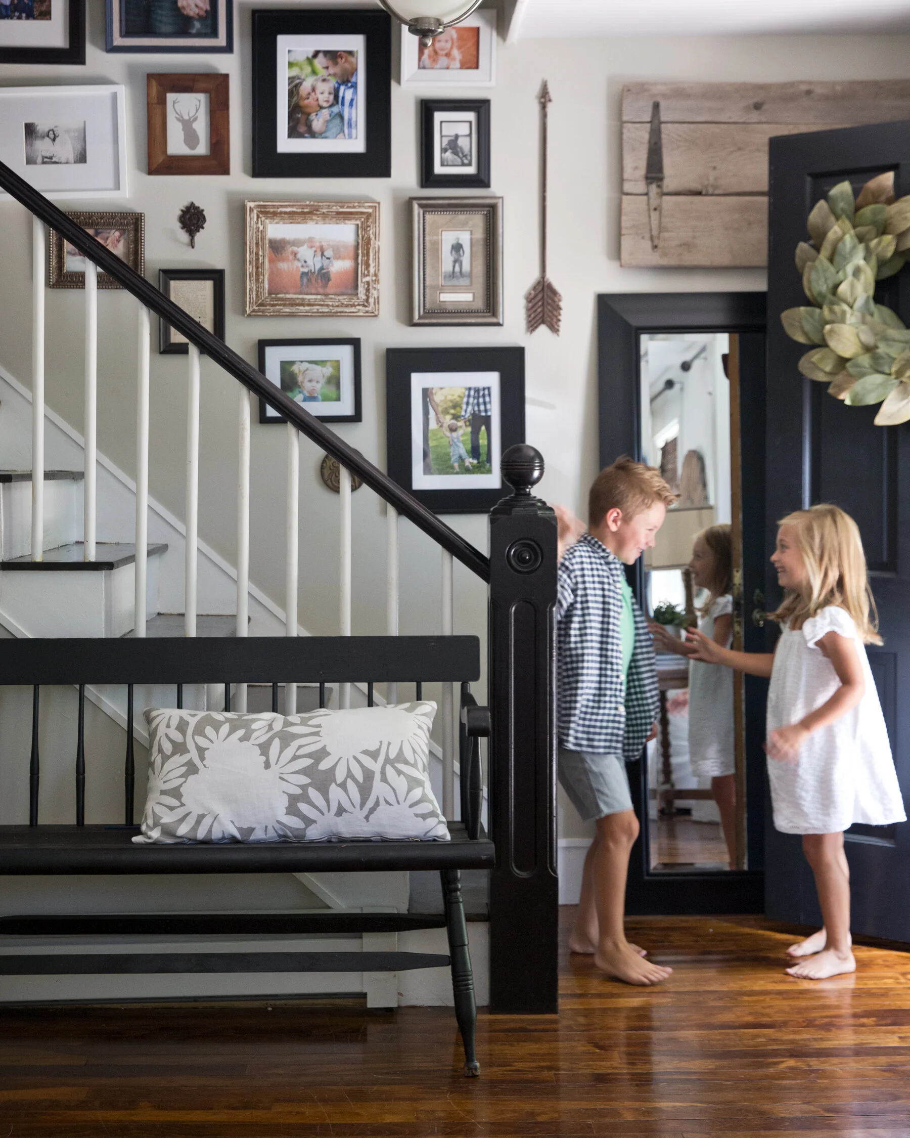 Three children, two girls and one boy, are standing near the door inside a home, smiling and talking. The wall behind them is decorated with a gallery of framed photos and artwork, and there's a black framed mirror on the door. A bench with a pattern