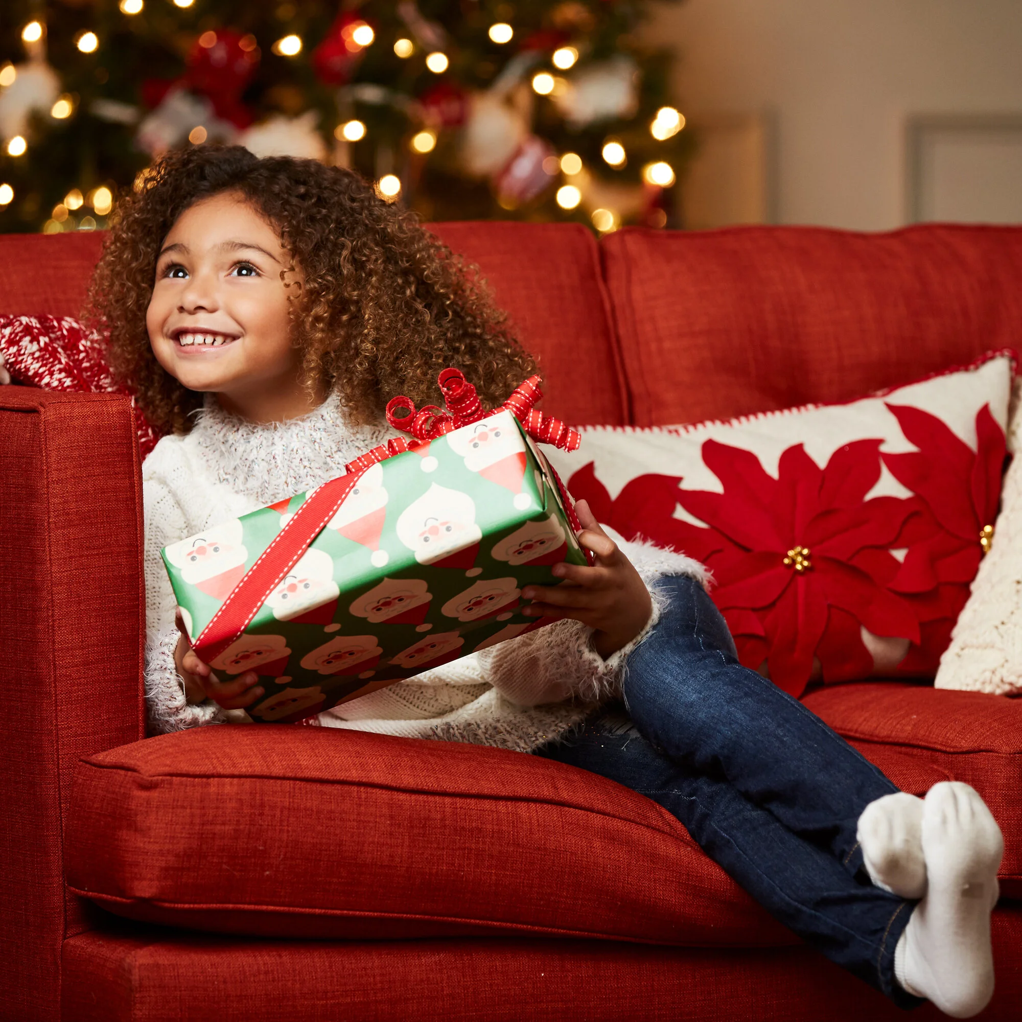 A young girl with curly hair smiling while sitting on a red sofa holding a Christmas gift wrapped with Santa Claus patterns, in front of a decorated Christmas tree.