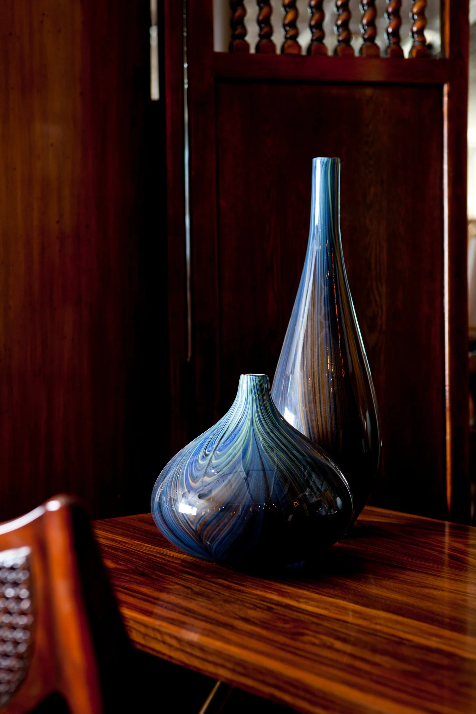 Two elongated glass vases with multicolored swirl patterns on a polished wooden table in a room with dark wood paneling.