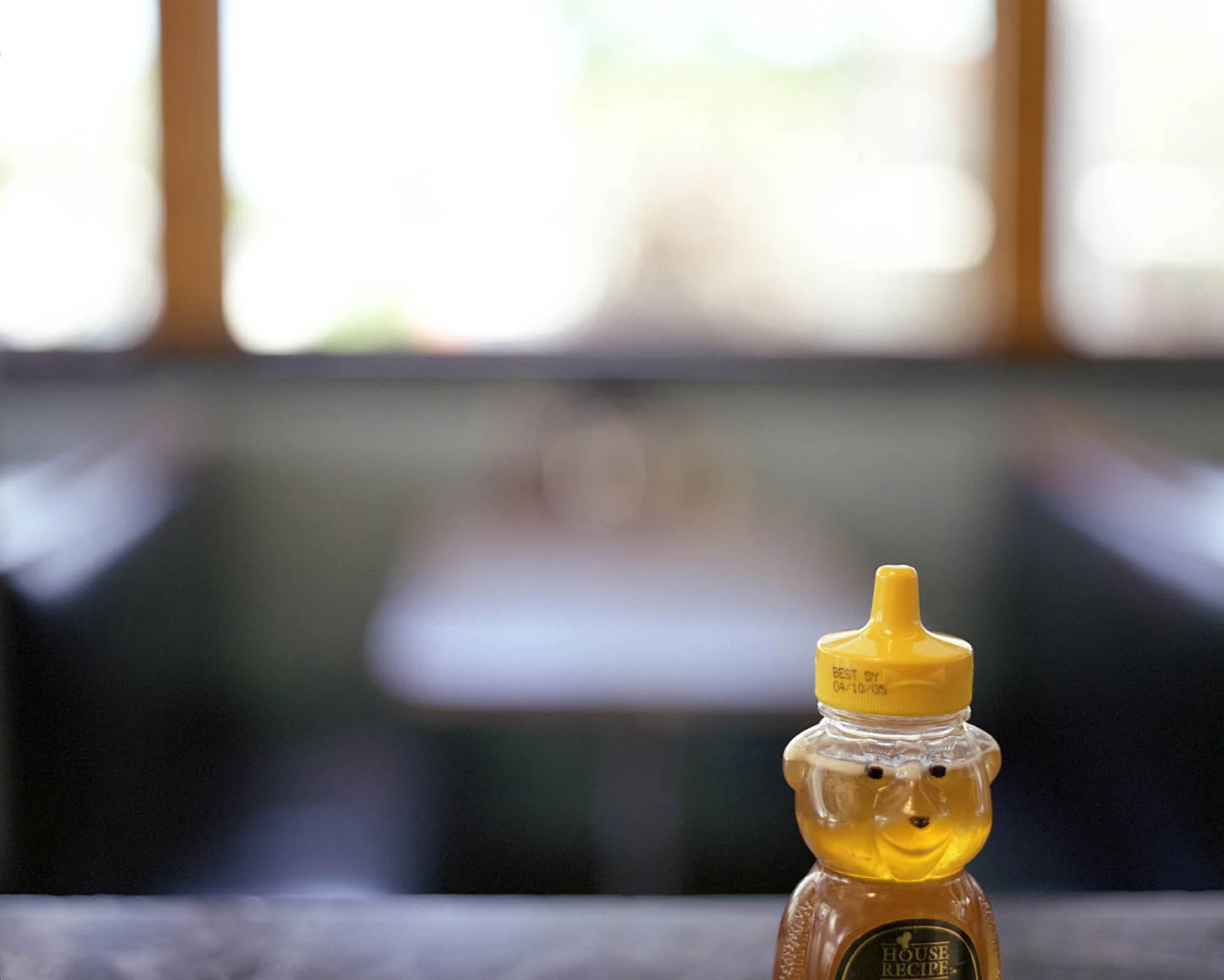 A honey bear syrup bottle with a honey-colored cap shaped like a bear's head on a kitchen counter with blurred background.