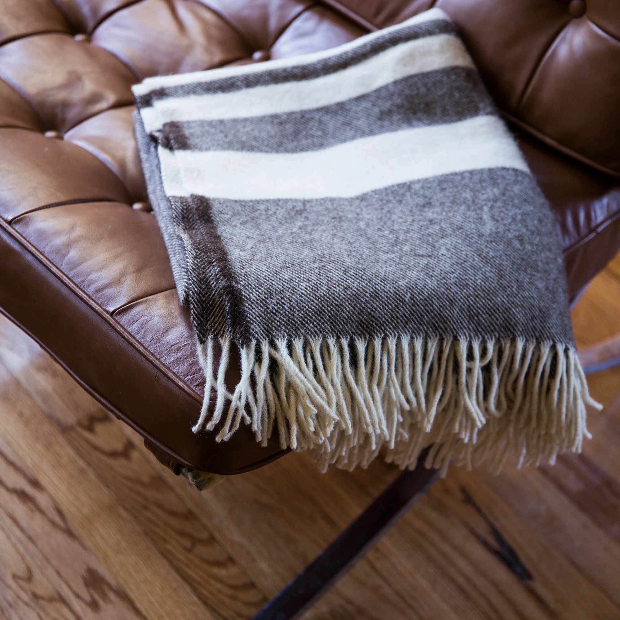 A brown leather armchair with a gray and white striped fringed blanket draped over it, sitting on a wooden floor.