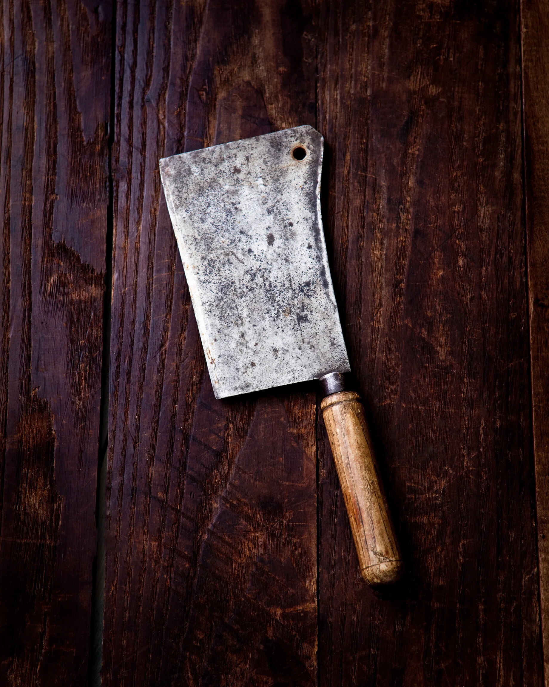 Old metal meat cleaver with wooden handle on dark wooden surface.