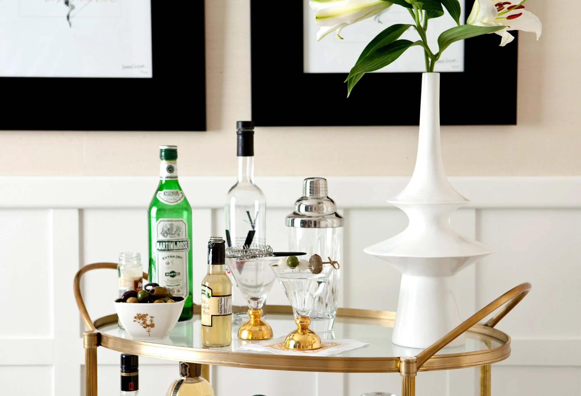 A gold bar cart with a white vase holding white lilies, two martini glasses with olives, various liquor bottles, and a bowl of mixed olives, with framed black and white botanical art in the background.