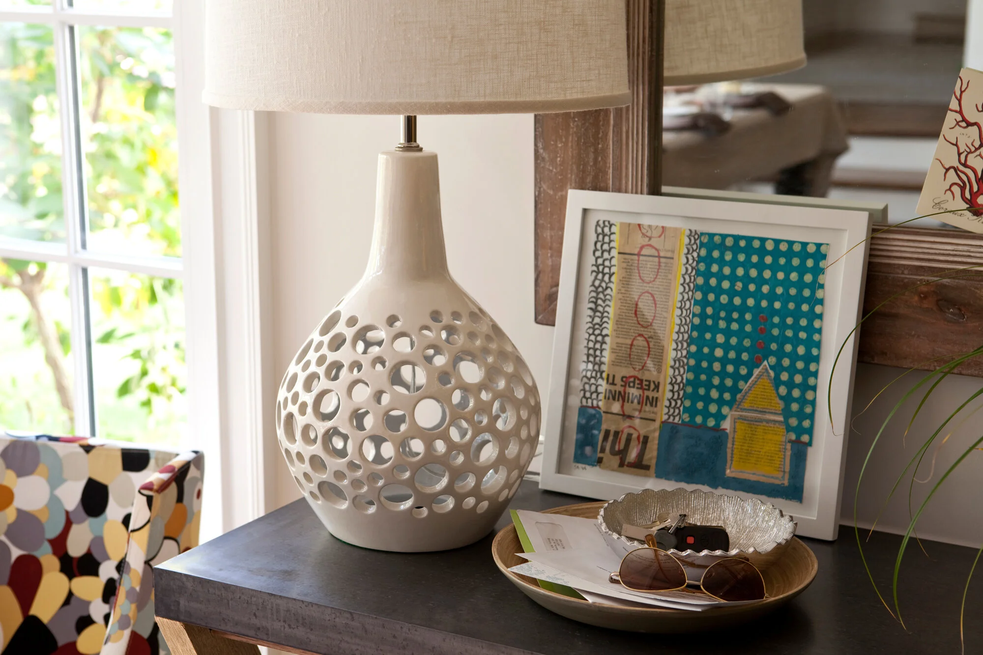 A white ceramic table lamp with a circular cutout design, placed on a dark wooden table, next to framed colorful artwork, sunglasses, and a silver dish with papers and keys. Window with natural light and greenery outside in the background.
