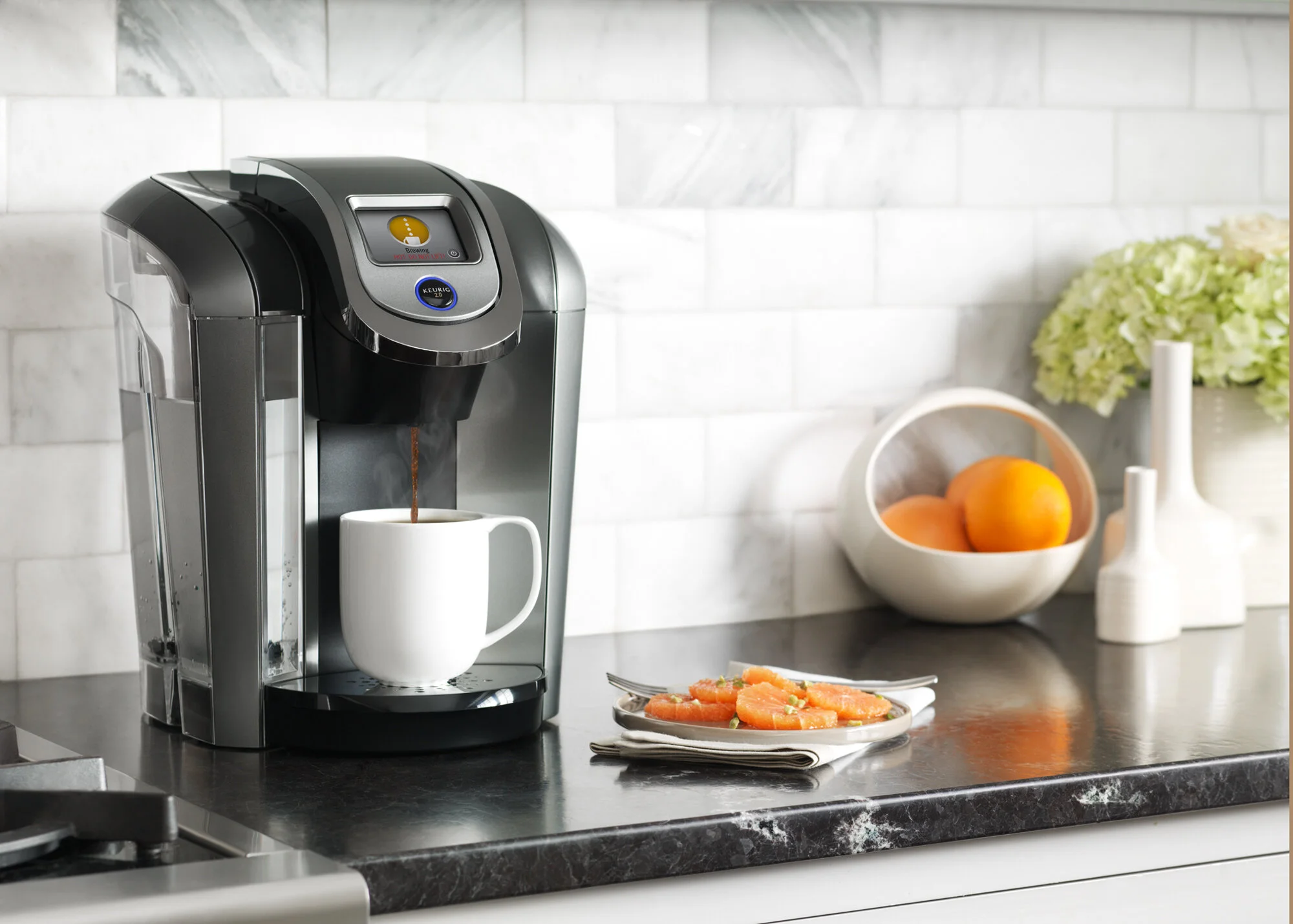 Keurig coffee maker brewing coffee into a white mug on a kitchen countertop. Next to it, a plate of sliced oranges, a bowl of fresh oranges, and a vase with white flowers are visible on the counter. Photographed by Joseph Keller for Keurig