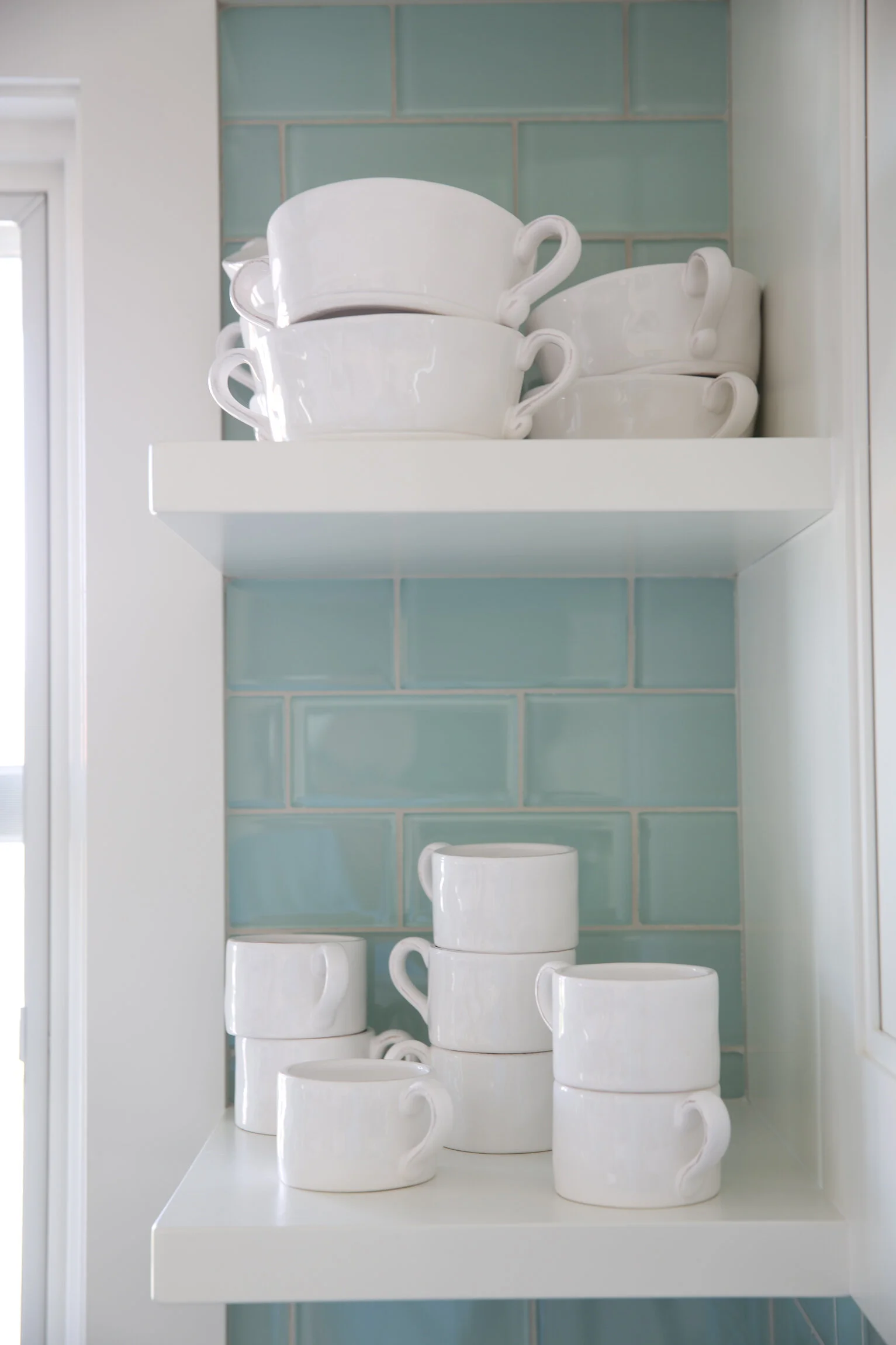 White cups and bowls stacked on kitchen shelves with blue tiled backsplash.